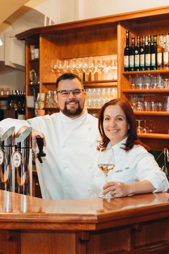 Ein Mann und eine Frau in weißen Kochjacken stehen hinter einer Schank aus Holz.