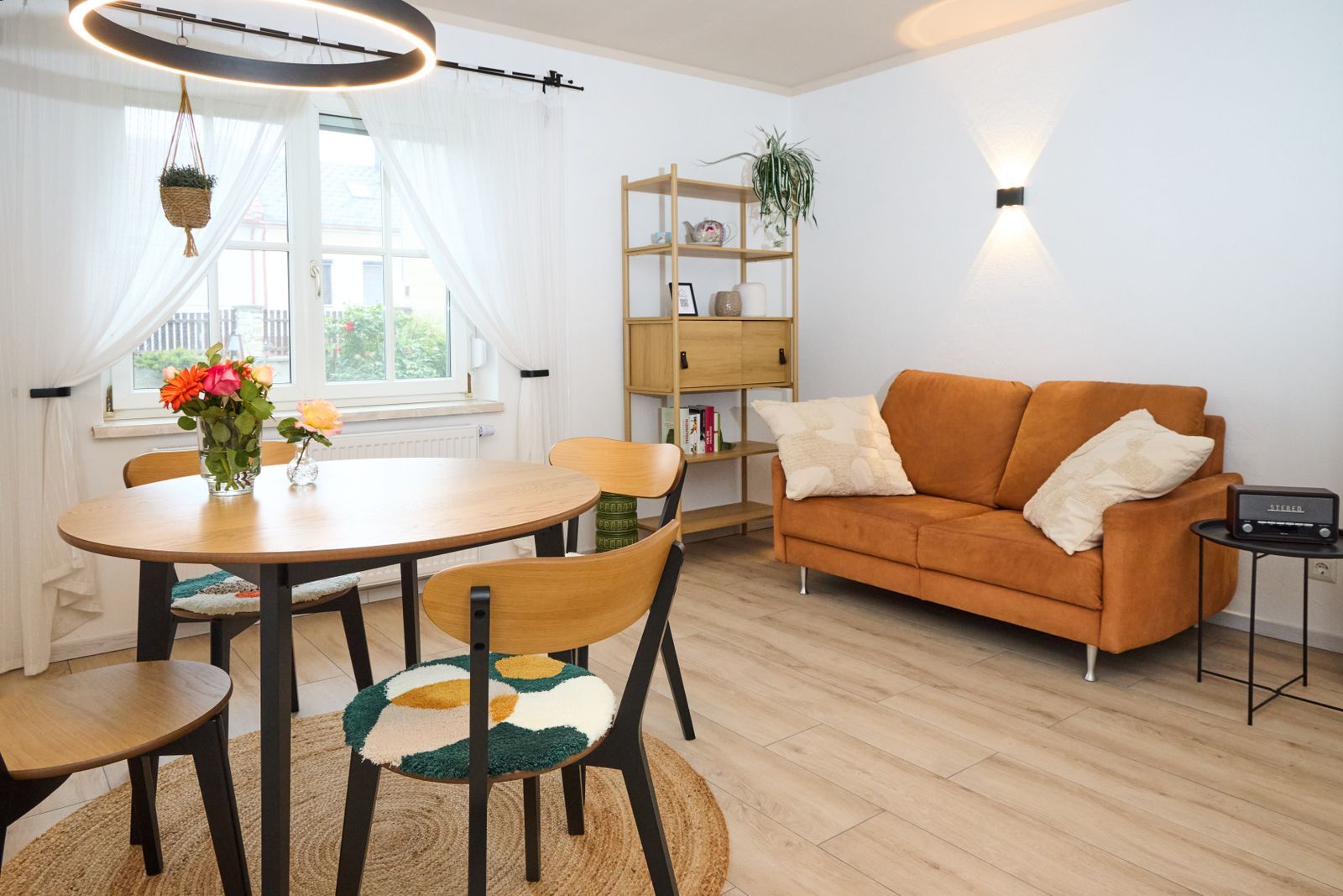Bright living and dining area with round dining table, four chairs, comfortable couch, shelving, large windows and modern lighting concept.