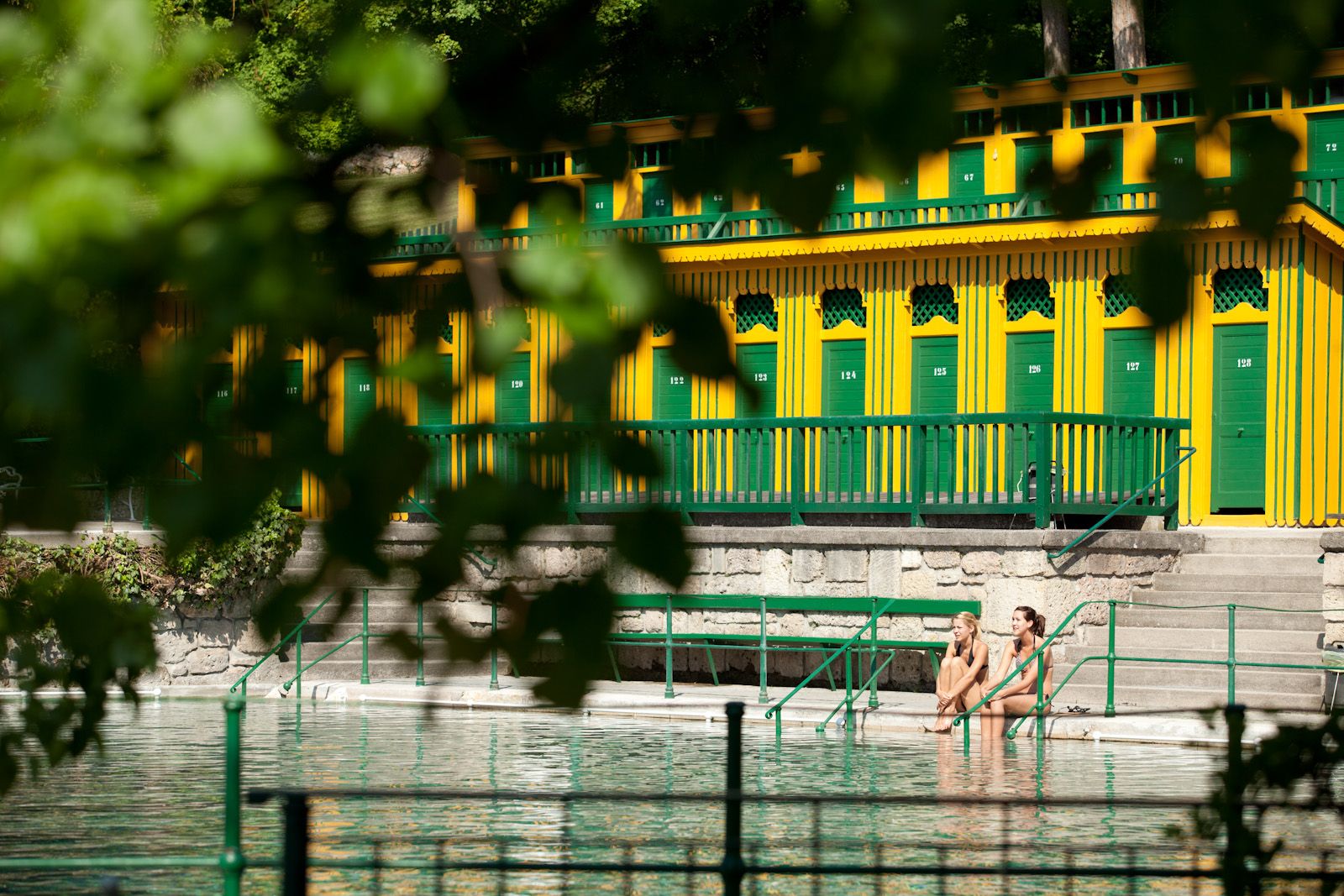 Two people sit on the edge of a thermal bath with yellow-green changing rooms in the background.