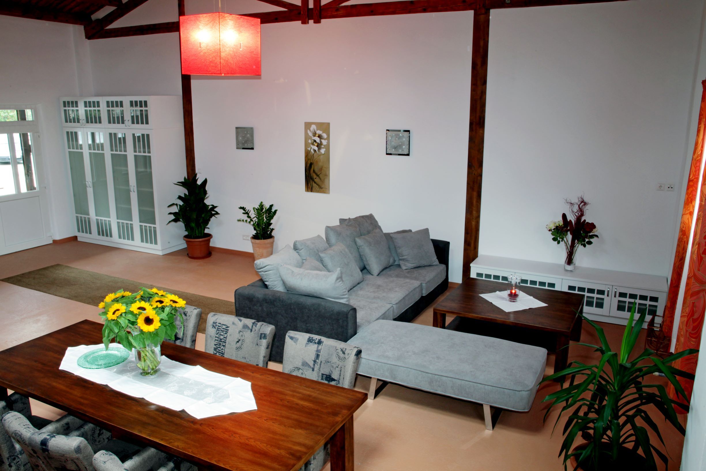 Ein modernes Loft mit grauem Sofa, Esstisch mit Sonnenblumen, Pflanzen und roten Lampenschirm.
