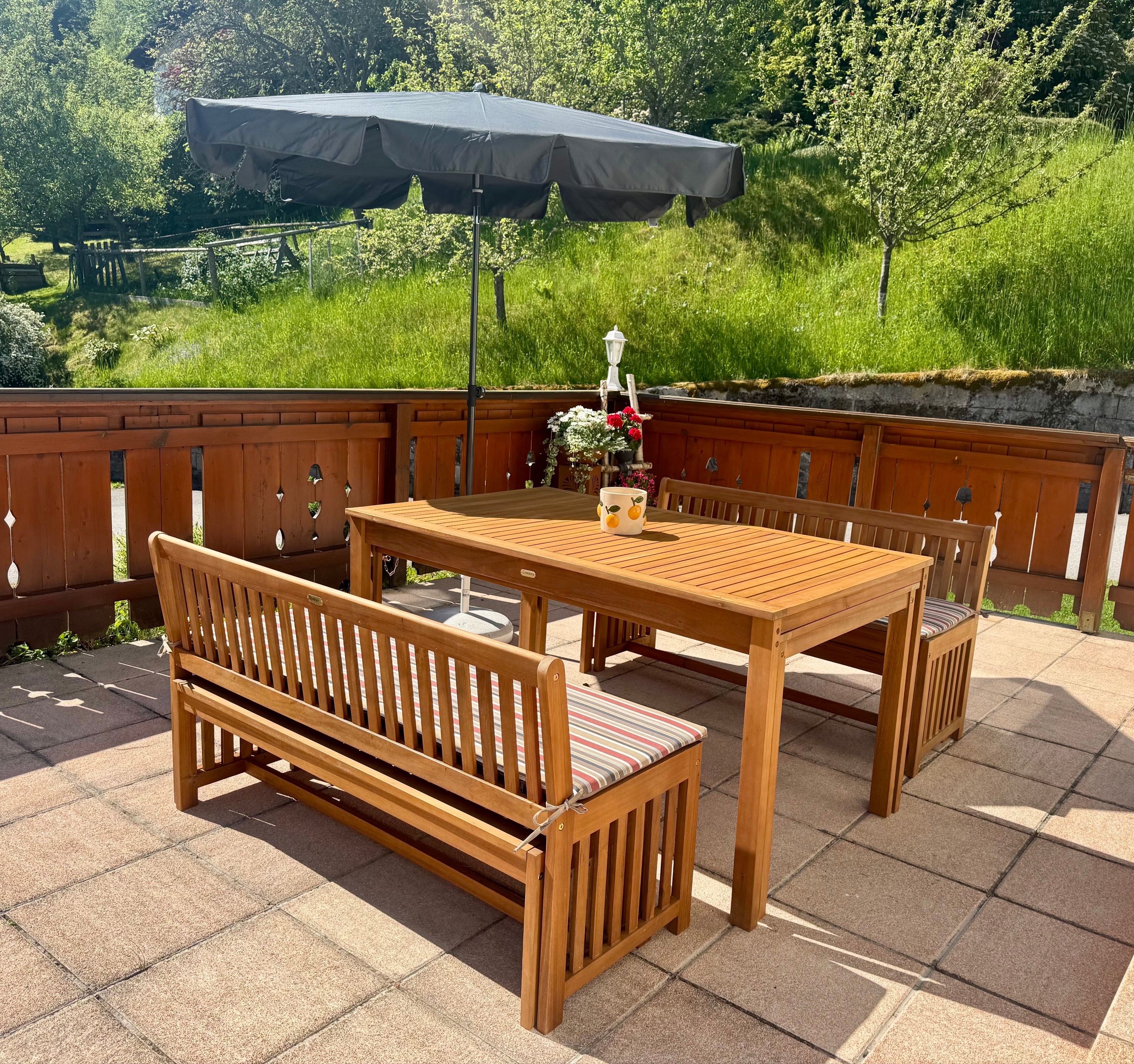Holzterrasse mit Tisch, Bänken und Sonnenschirm, umgeben von grünem Garten.