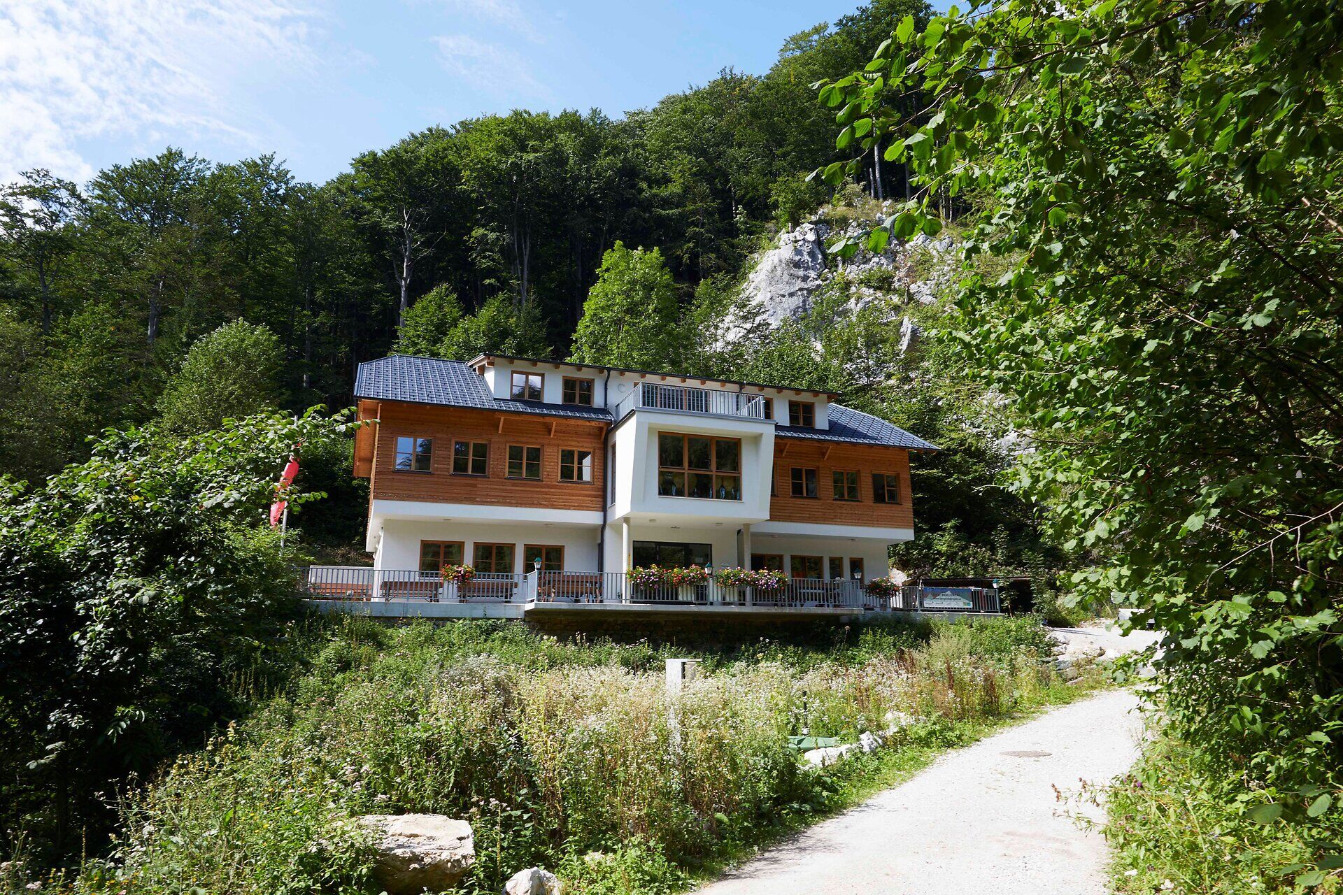 Hütte liegt zwischen vielen Bäumen am Beginn einer Felsschlucht, im Vordergrund ist ein Weg.