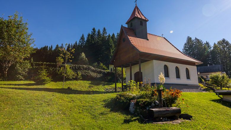 Kleine Kapelle mit rotem Dach auf einer grünen Wiese, umgeben von Bäumen und Blumen.