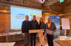Vier Personen stehen in einem rustikalen Raum vor einer Leinwand nebeneinander und halten ein Wiener Alpen Schild sowie ein LEADER-Schild in den Händen. 