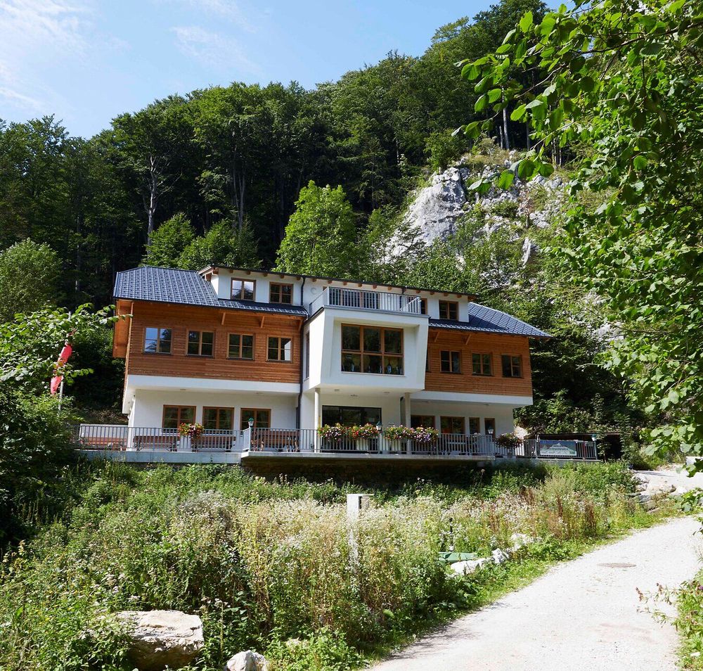 Hütte liegt zwischen vielen Bäumen am Beginn einer Felsschlucht, im Vordergrund ist ein Weg.