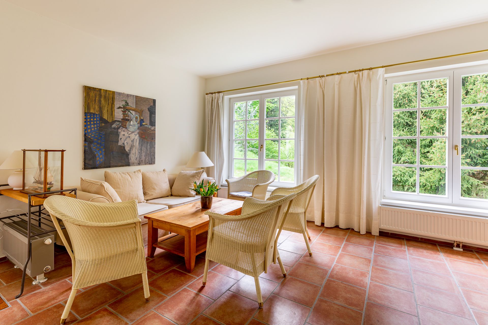 Heller Wohnraum mit Sofa, Korbstühlen, Holztisch und großen Fenstern mit Blick ins Grüne.