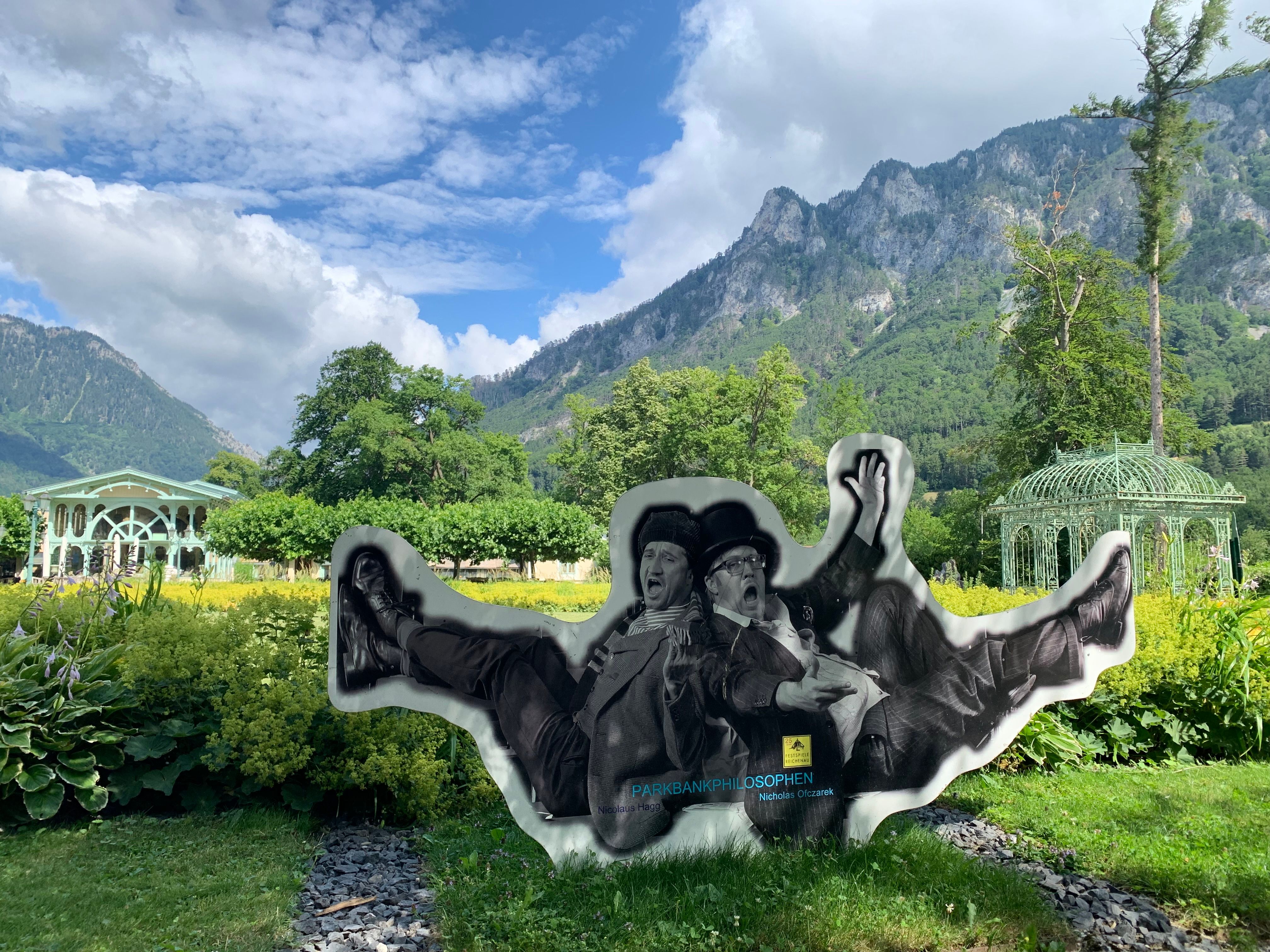 Eine Gartenlandschaft mit Bergen im Hintergrund und einer Skulptur von zwei sitzenden Personen im Vordergrund.