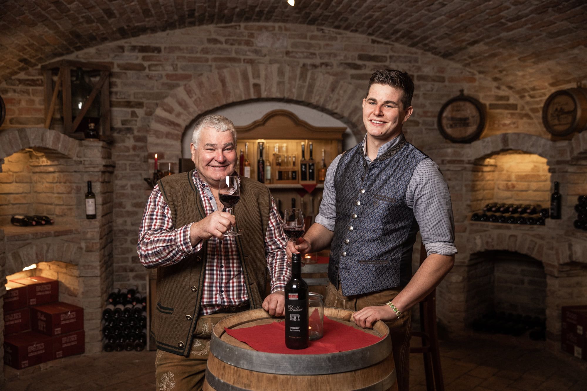 Ein junger und ein älterer Mann in einem Weinkeller mit Weingläsern in der Hand vor einem Holzfass.