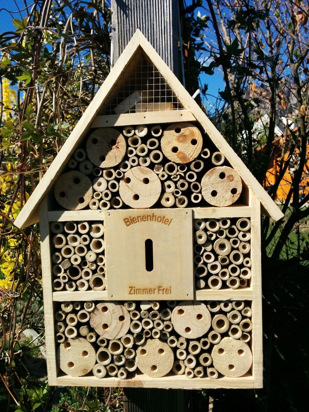 Ein Bienenhotel aus Holz mit verschiedenen Röhren und Löchern, aufgehängt an einem Pfosten im Garten.