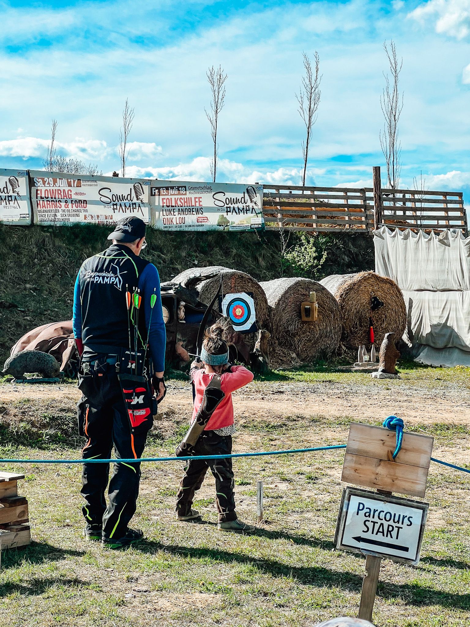 An adult and a child are standing on an archery course. In the background are targets and a sign with the words 'Parcours START'.