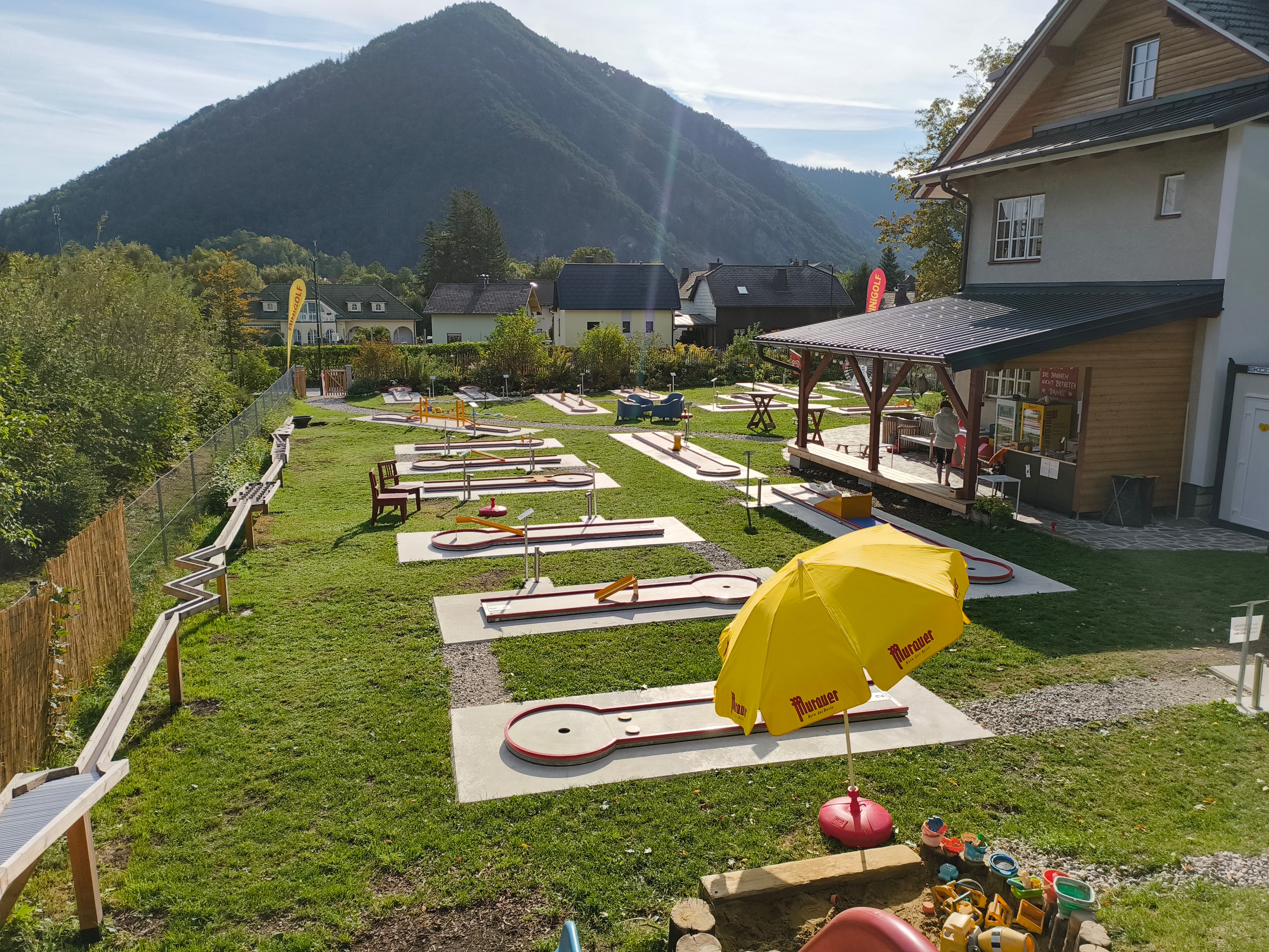 Minigolfplatz mit mehreren Bahnen und einem gelben Sonnenschirm, umgeben von Bergen und Häusern.