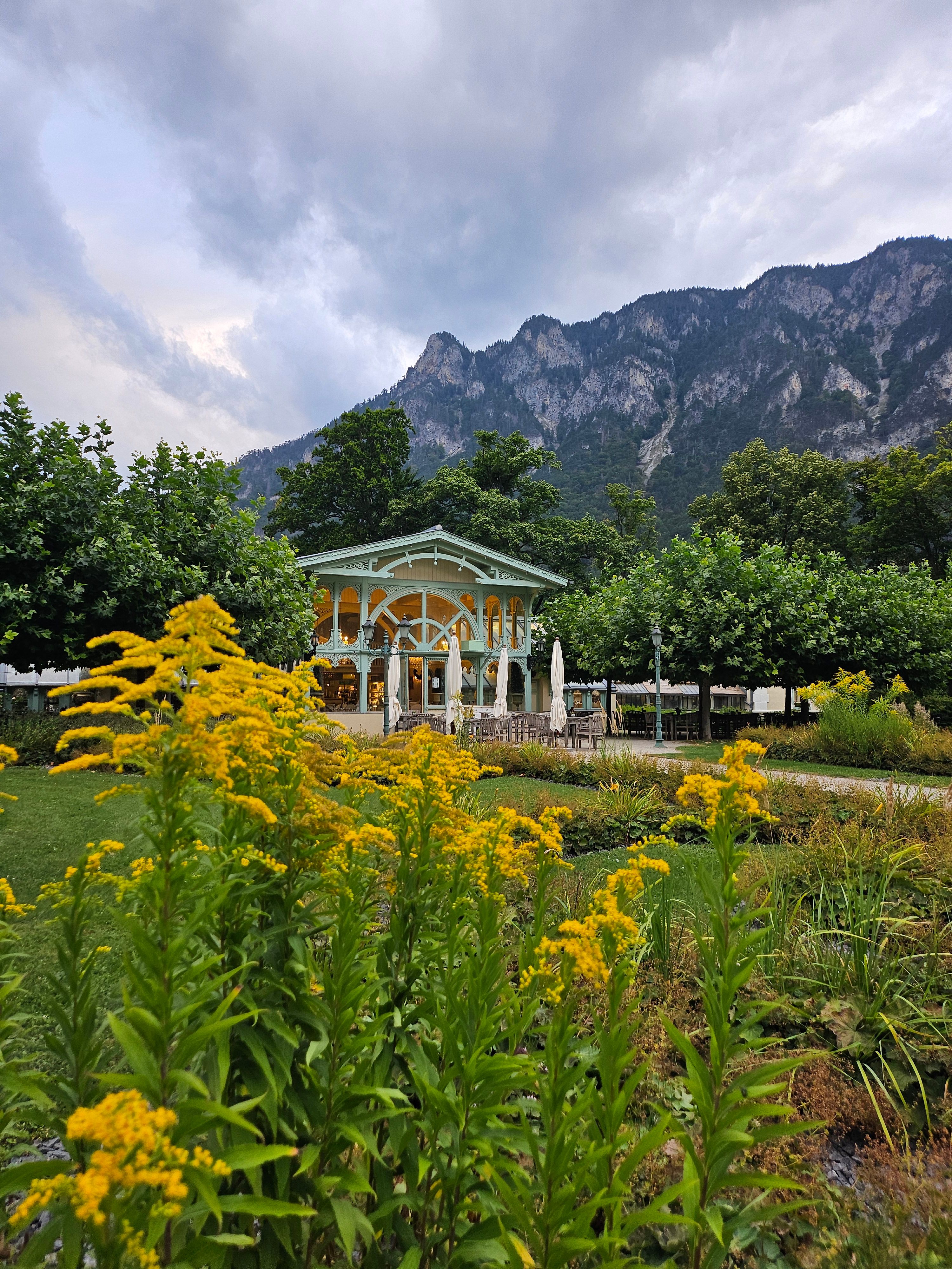 Ein Pavillon in einem Garten mit gelben Blumen im Vordergrund und Bergen im Hintergrund.
