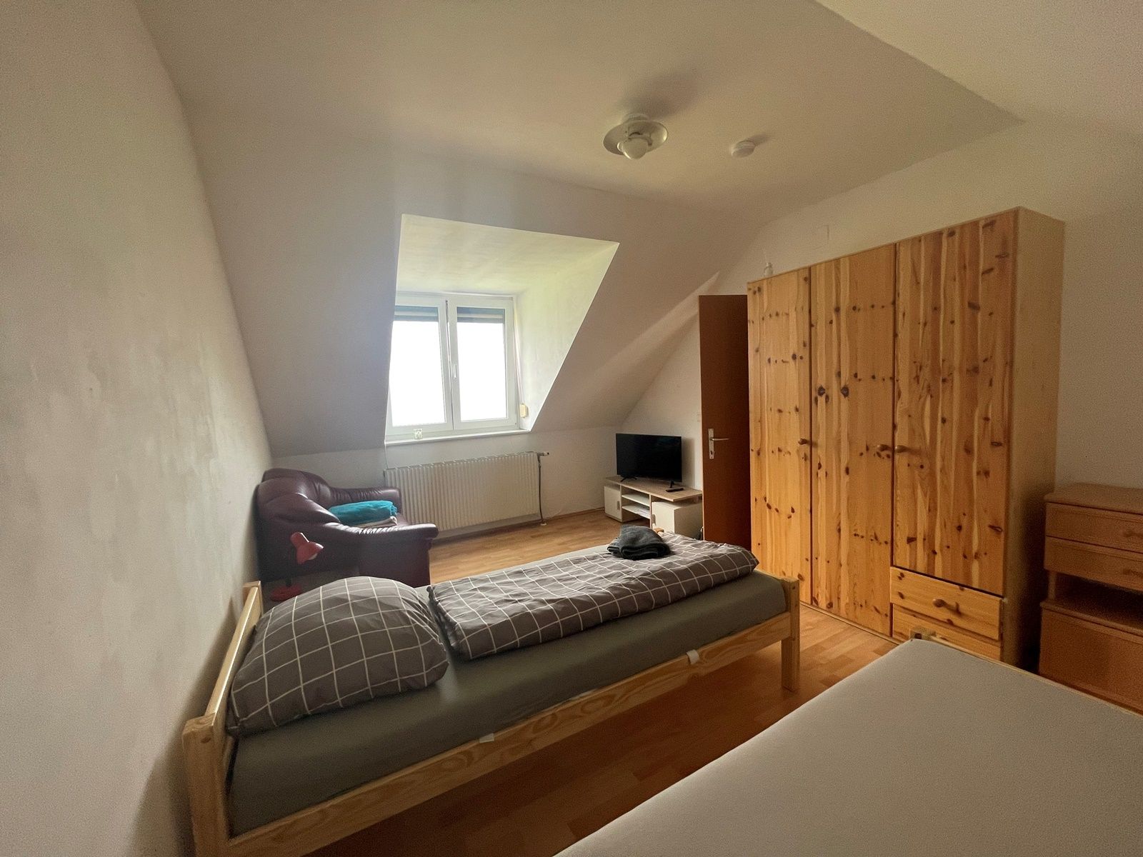 Ein einfach eingerichtetes Schlafzimmer mit zwei Betten, einem Kleiderschrank, einem Fernseher und einem Sessel unter einer Dachschräge.