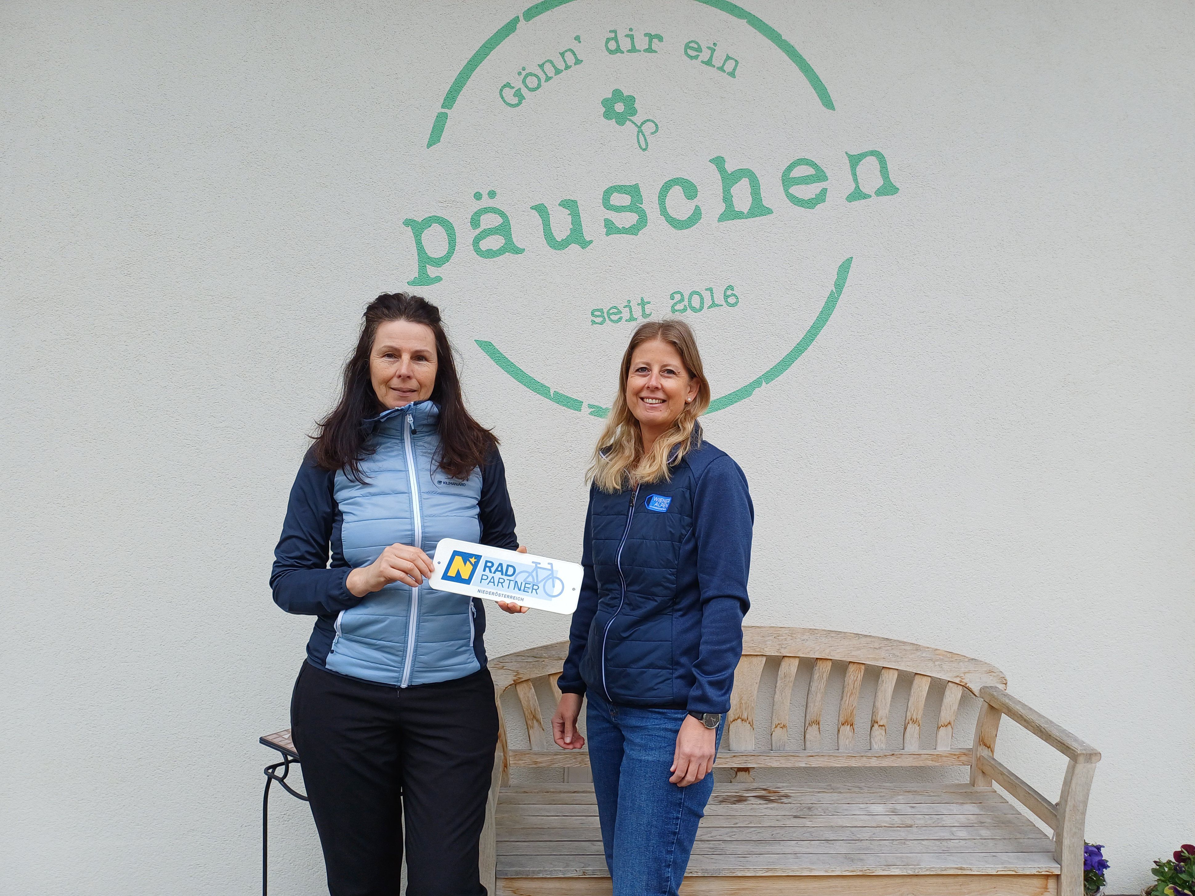 Zwei Frauen stehen vor einer Hauswand mit der Aufschrift "Päuschen". Eine Frau hält ein Radpartner Niederösterreich Schild in den Händen.
