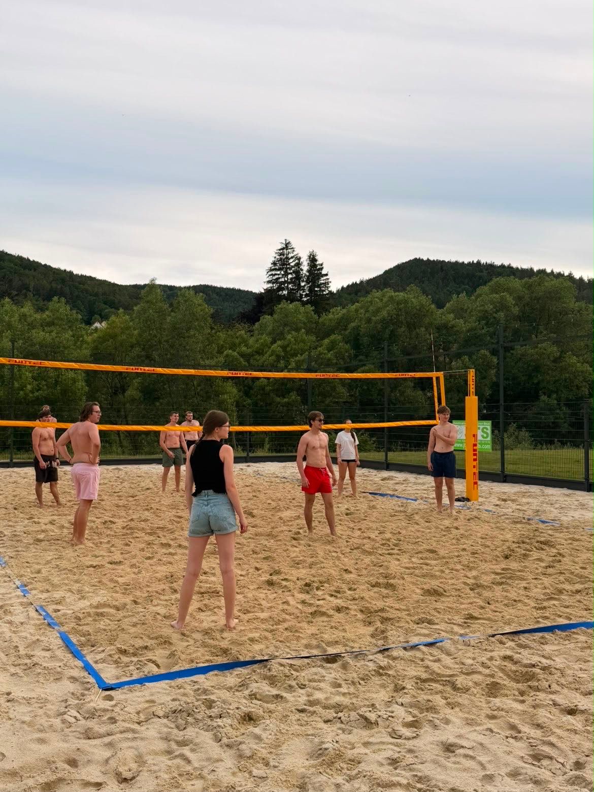 Group of people playing beach volleyball on an outdoor sand court surrounded by trees and hills.