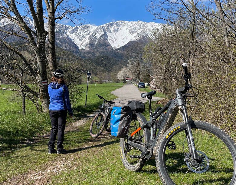 A person wearing a helmet stands next to bicycles on a path with snow-covered mountains in the background.
