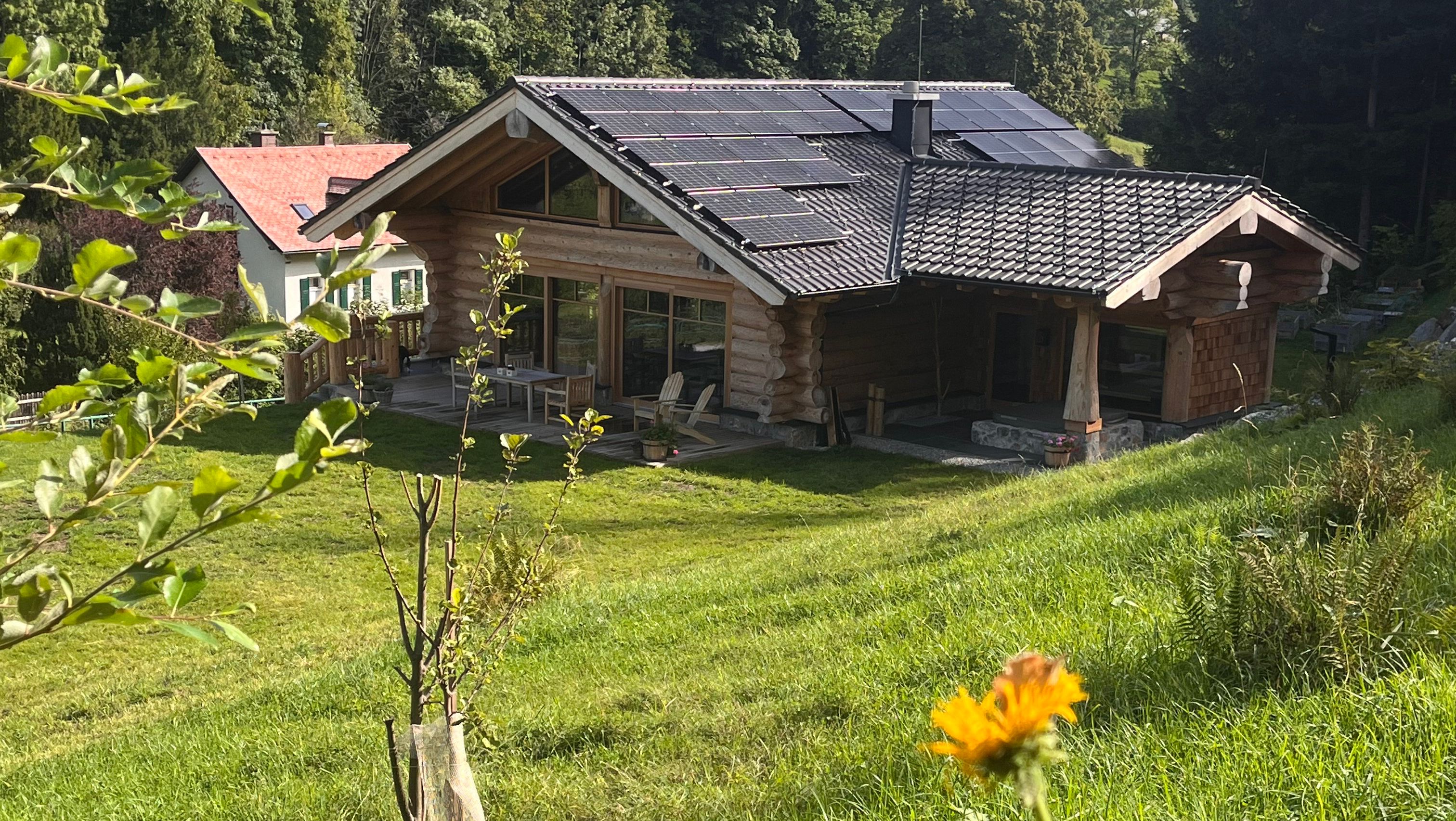 Ein Holzhaus mit Solarpanelen auf dem Dach, umgeben von grüner Landschaft und Bäumen.