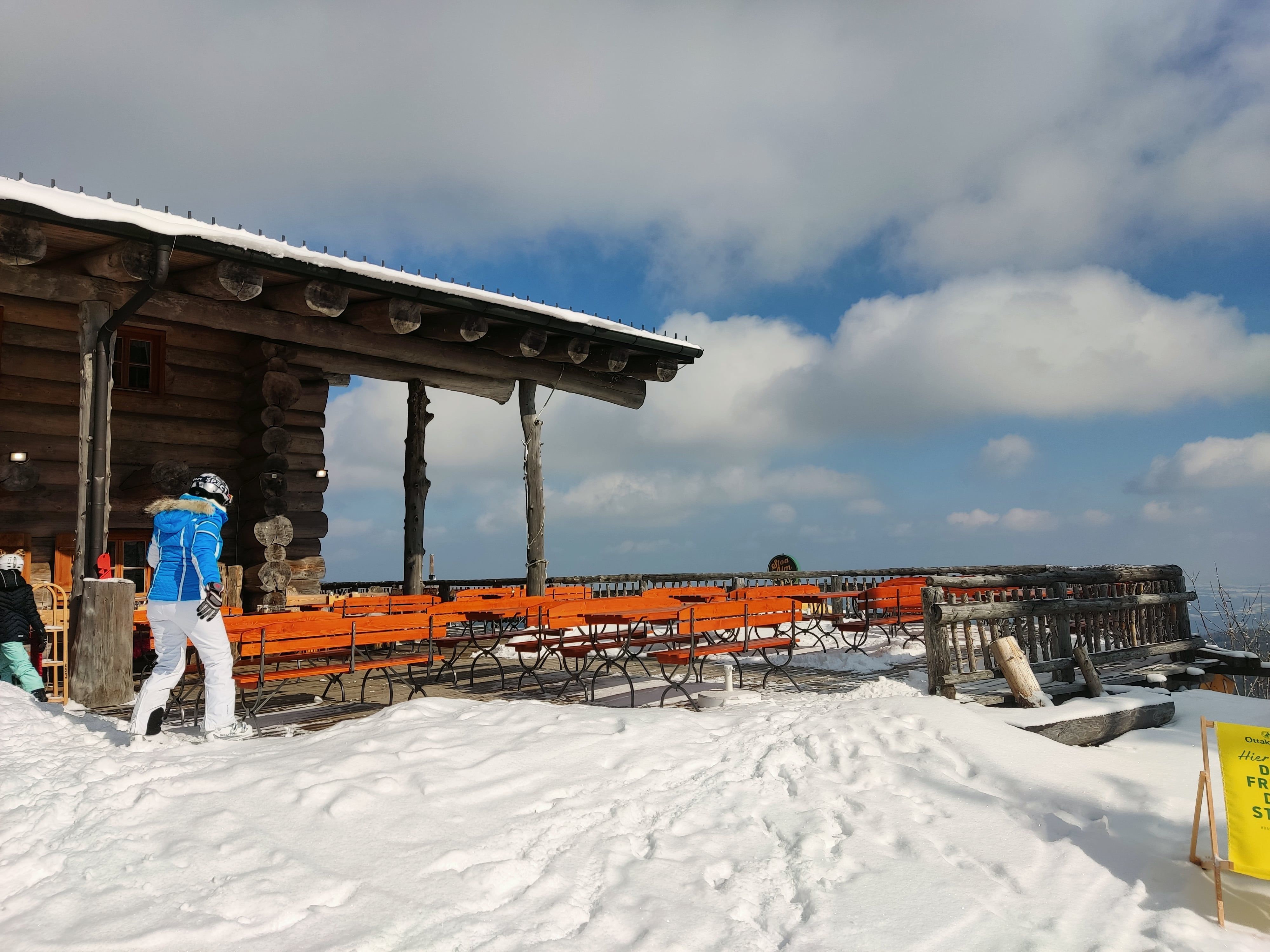 Verschneite Terrasse der Stoa Alm mit Holzbänken und Tischen, blauer Himmel und Wolken im Hintergrund.
