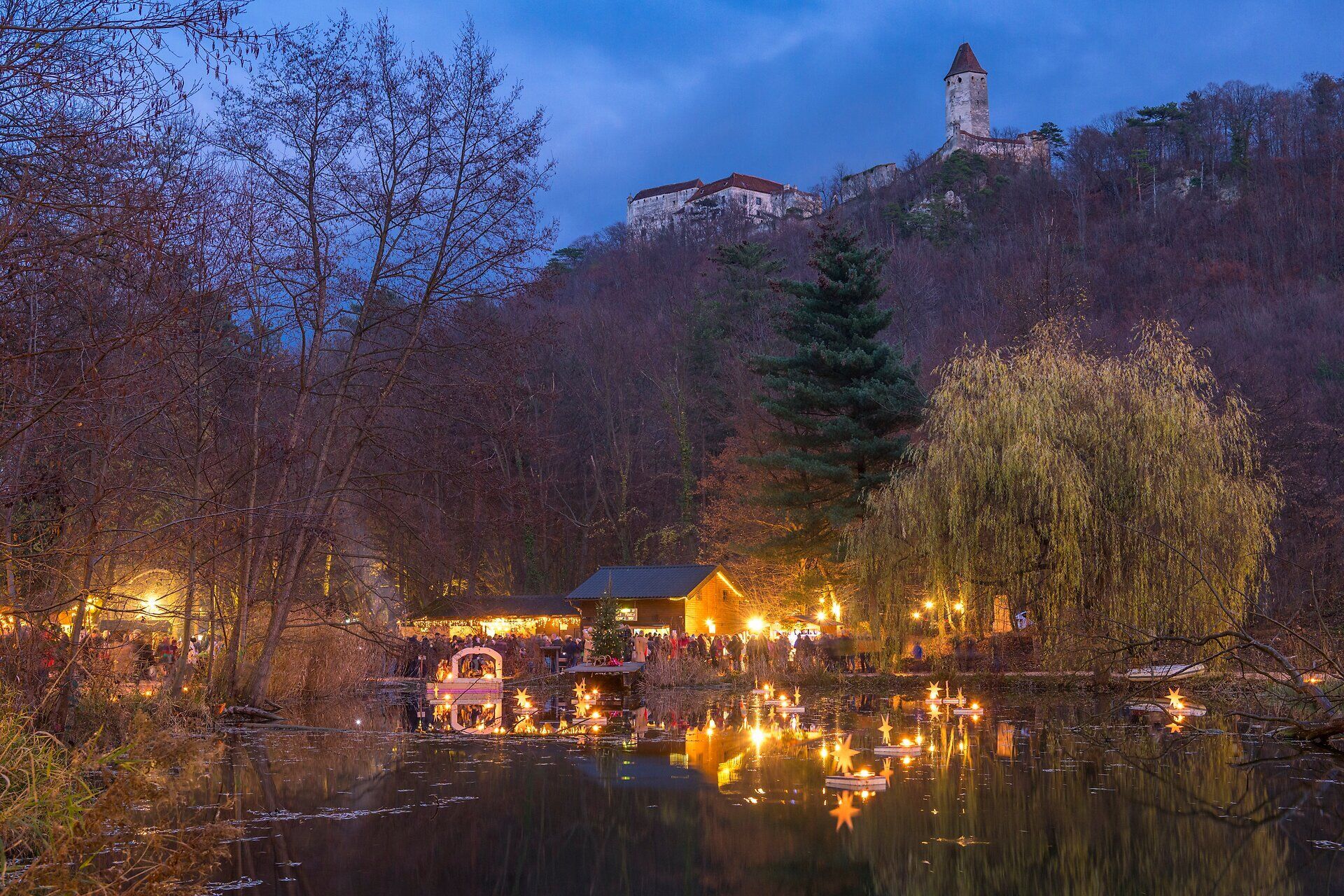 Beleuchteter Weihnachtsmarkt am Teich mit Spiegelungen im Wasser und Burg im Hintergrund bei Dämmerung.