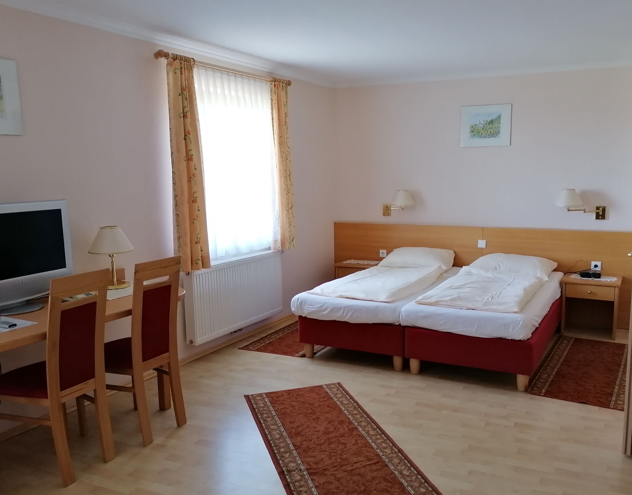 Ein Doppelzimmer mit zwei Einzelbetten, einem Schreibtisch mit Stühlen, einem Fernseher und einem Fenster mit Vorhängen.