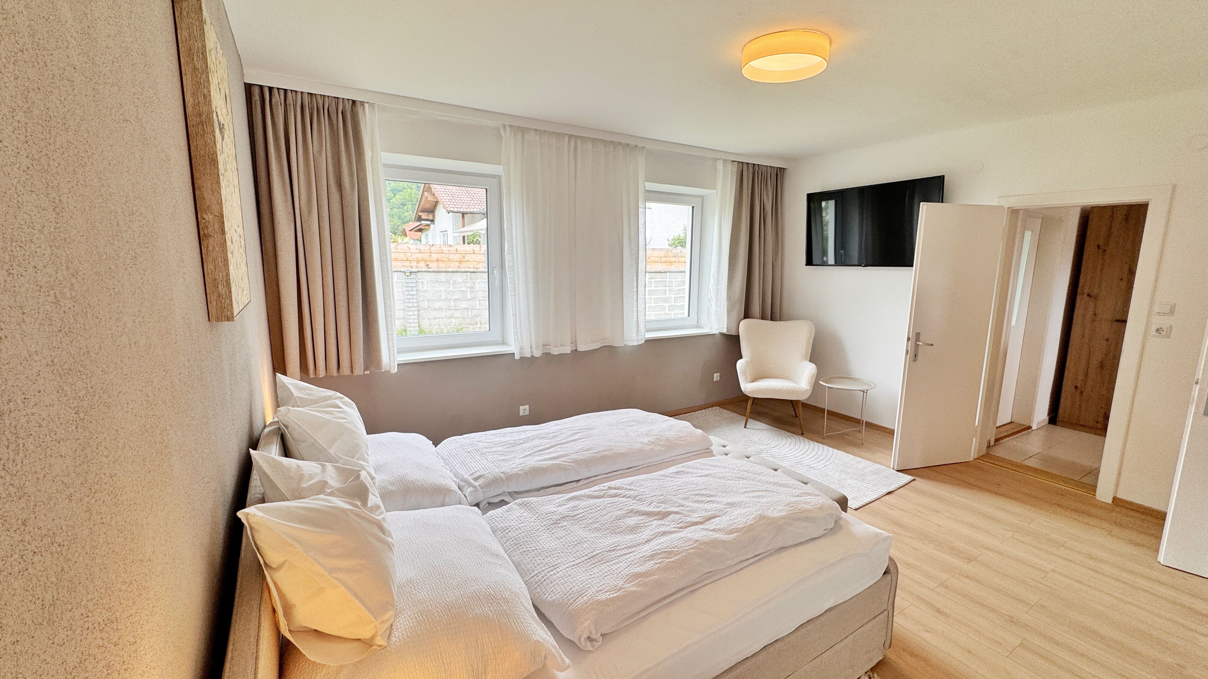 Modernes Schlafzimmer mit Doppelbett, zwei Fenstern, Sessel und Fernseher an der Wand.