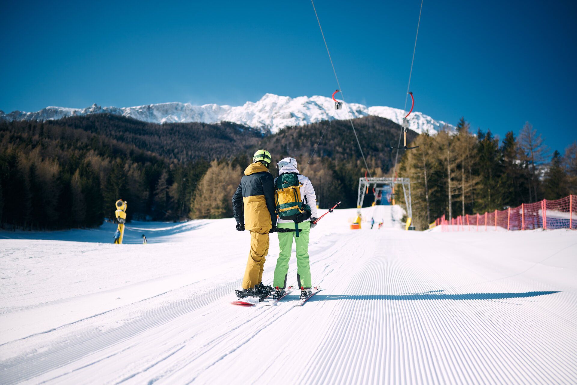 Zwei Skifahrer stehen auf der perfekt präparierten Piste und genießen die atemberaubende Aussicht auf die schneebedeckten Berge. Die klare, kalte Luft und die strahlende Sonne schaffen eine unvergessliche Winteratmosphäre, die zum Verweilen einlädt.