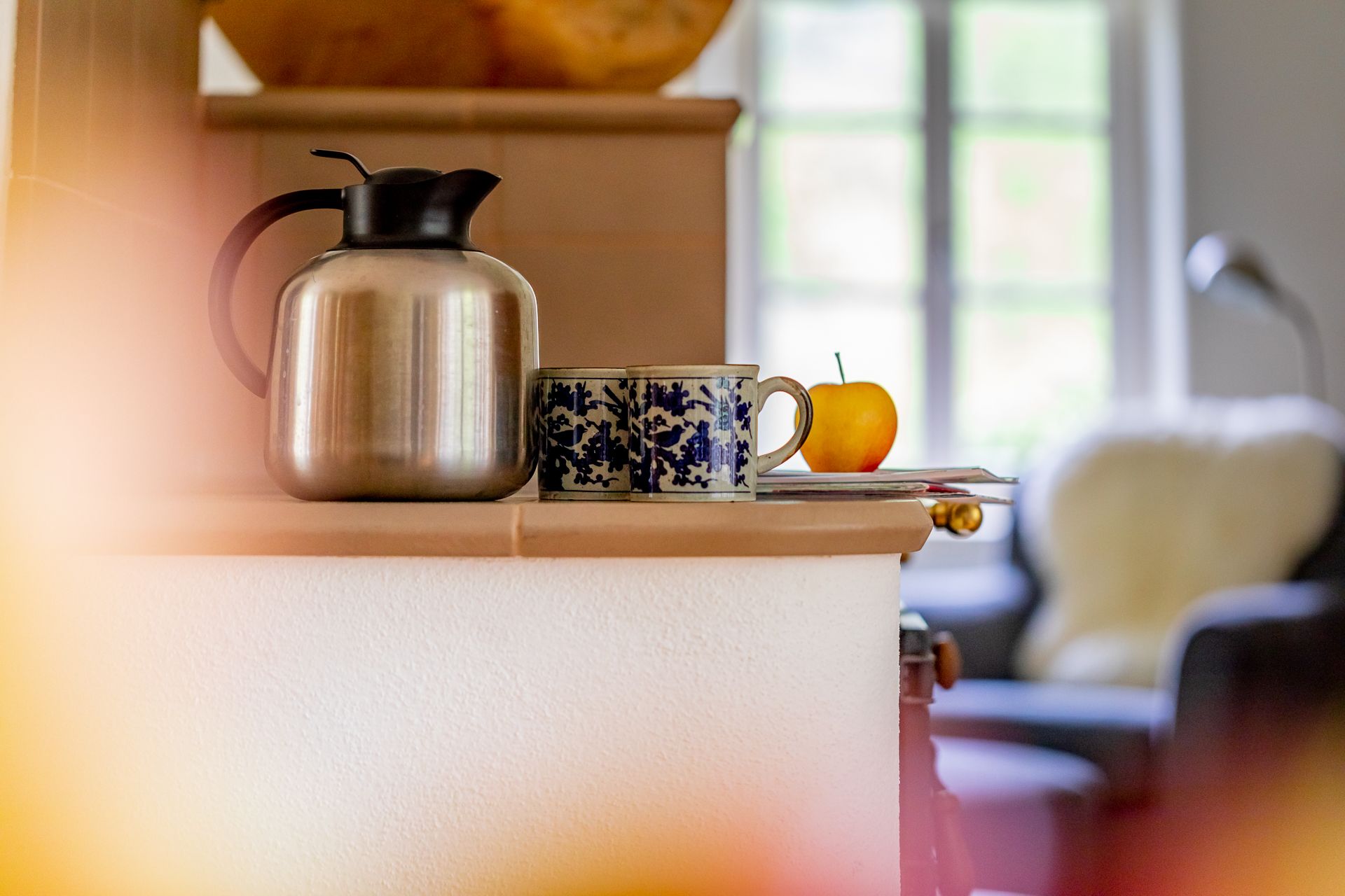 Ein Tisch mit einer Thermoskanne, zwei Tassen und einem Apfel vor einem Fenster.