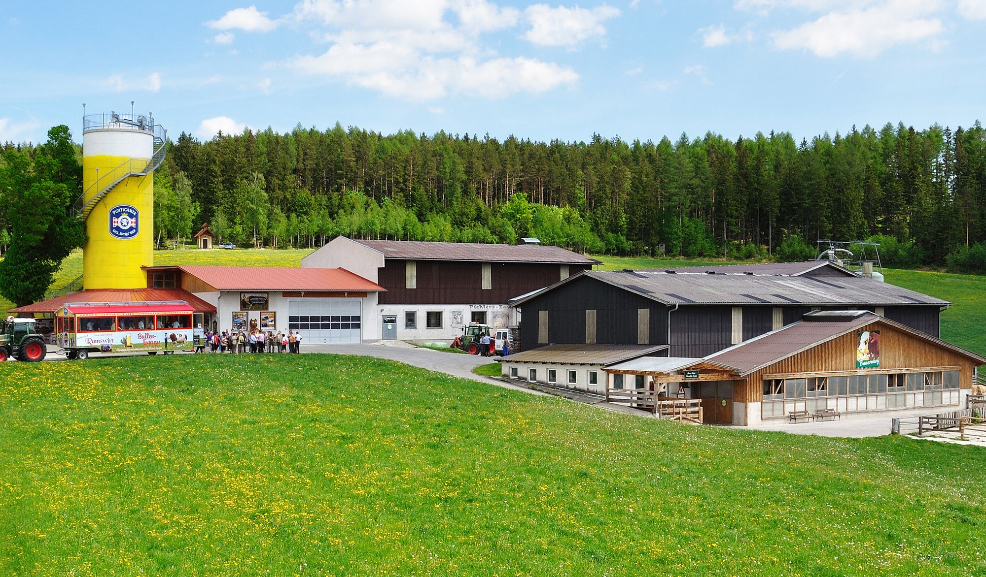 Bauernhof mit gelbem Silo und Traktoranhänger vor grüner Wiese.