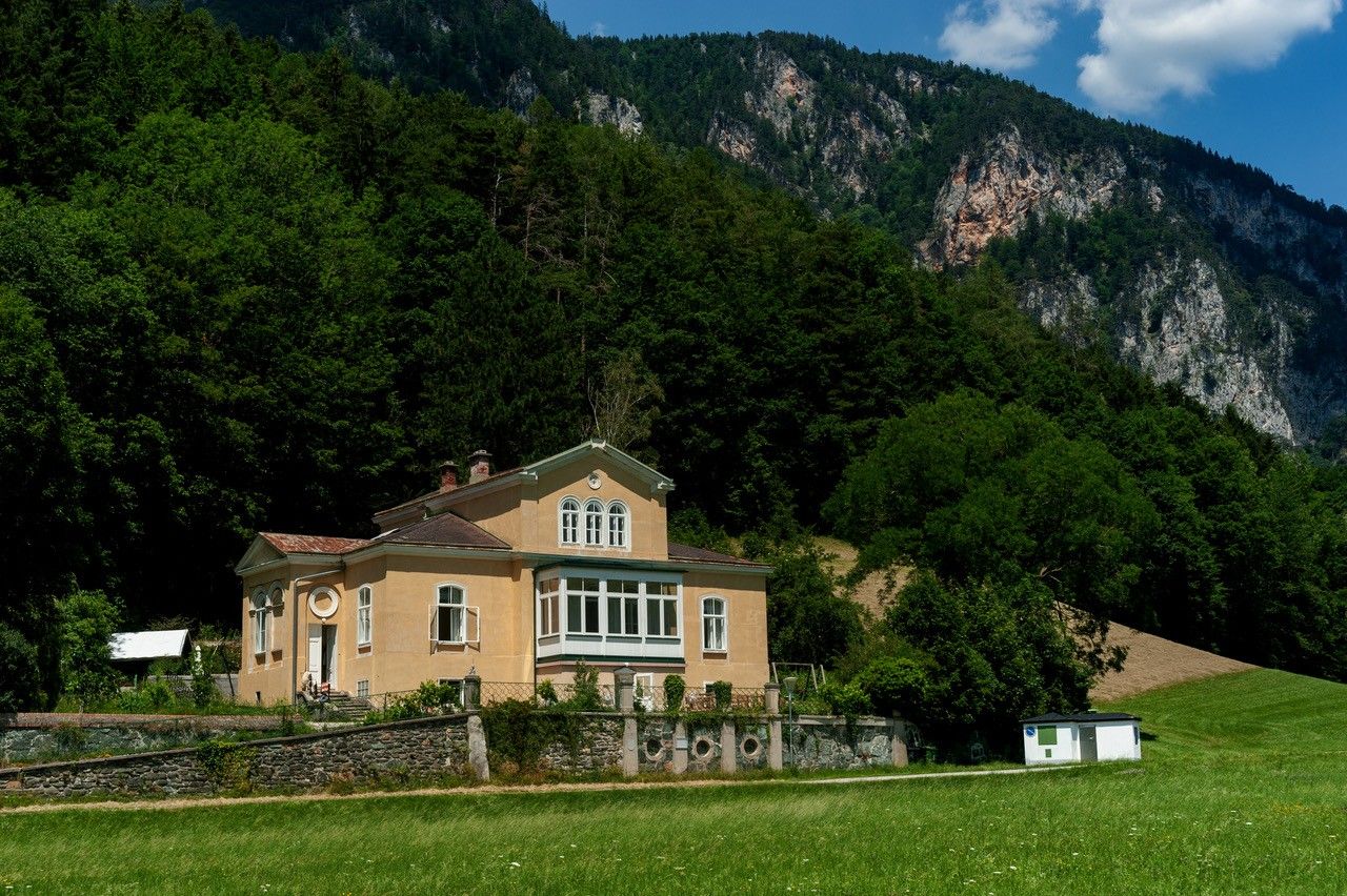 Ein gelbes Haus mit großen Fenstern steht vor einem bewaldeten Berghang unter blauem Himmel.