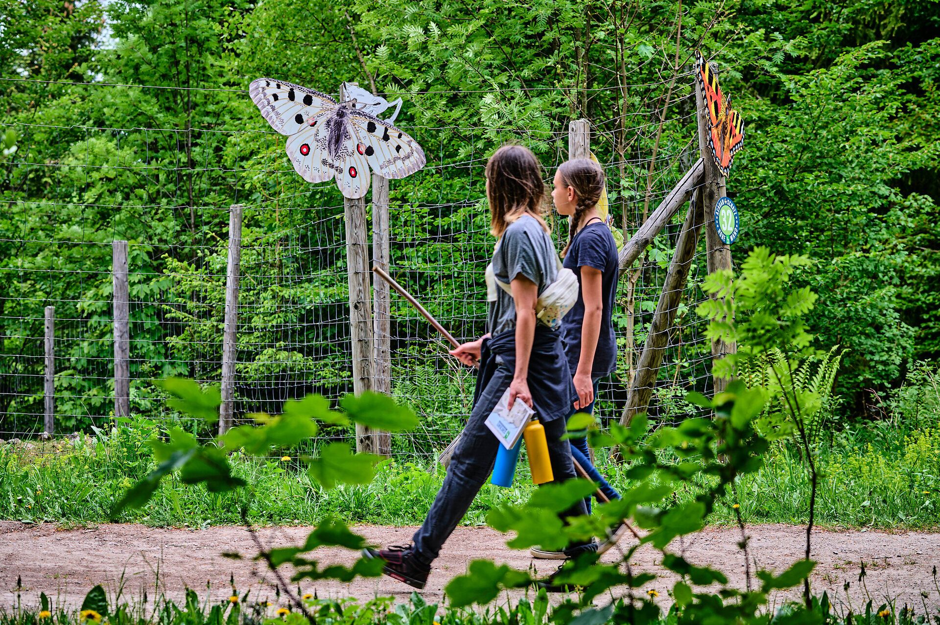 Zwei Kinder schlendern fröhlich auf einem schmalen Weg, umgeben von üppigem Grün und bunten Schmetterlingen, die an den kunstvoll gestalteten Schildern vorbeifliegen. Die frische Luft und die lebendige Natur schaffen eine einladende Atmosphäre, die zum Entdecken und Verweilen einlädt.