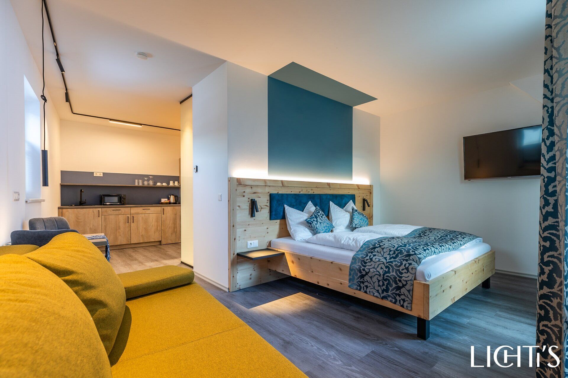 Das Zimmer im Lchti’s Rooms & Appartements ist ausgestattet mit einem Doppelbett, Sitzmöglichkeiten und Fernseher an der Wand. Dahinter befindet sich in einem abgetrennten Bereich eine Küchenzeile.