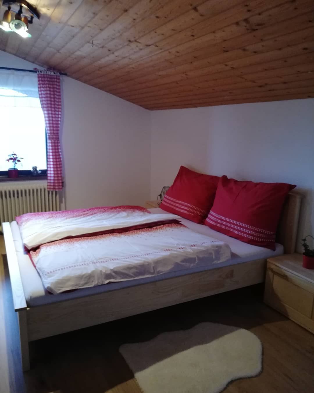 Gemütliches Schlafzimmer mit Holzbett, roten Kissen und karierten Vorhängen unter einer Holzdecke.