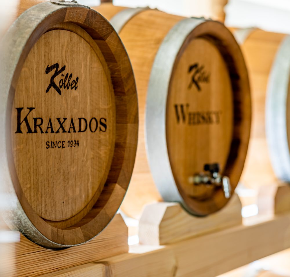 Zwei Holzfässer mit Kraxados und Whisky liegen auf einer Holzaufbarrung, auf den Vorderseiten ist der Name der Brennerei Kölbel, die Bezeichnung des Inhaltes und since 1994 gebrannt.