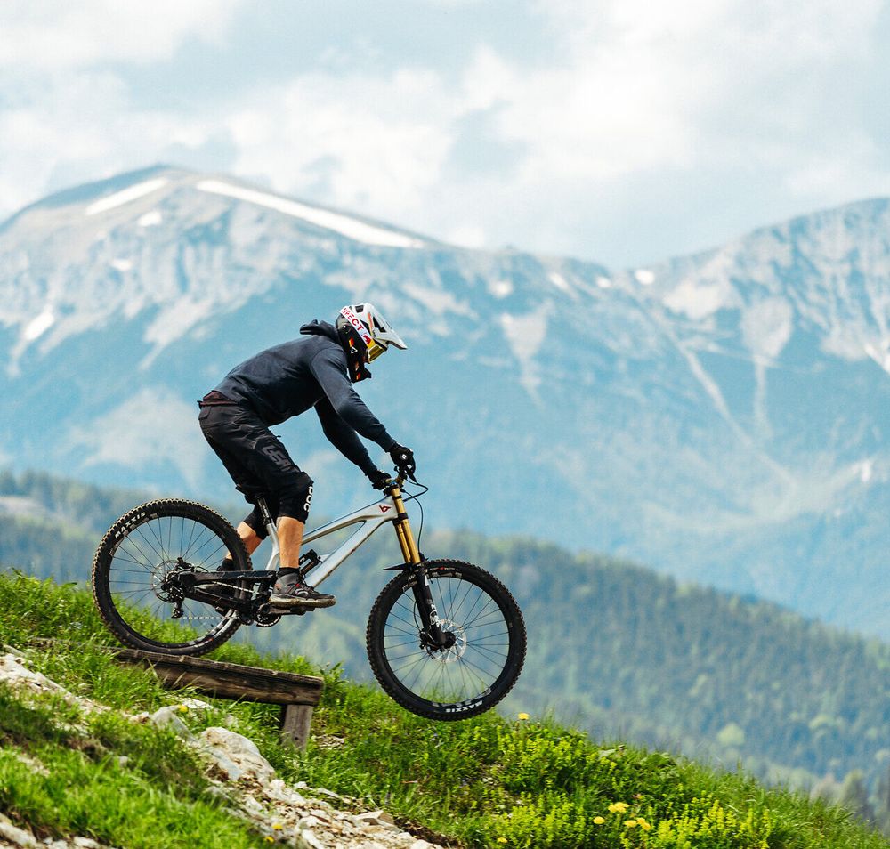 Mountainbiker fährt auf einem schmalen Trail durch alpine Landschaft mit Bergen im Hintergrund.