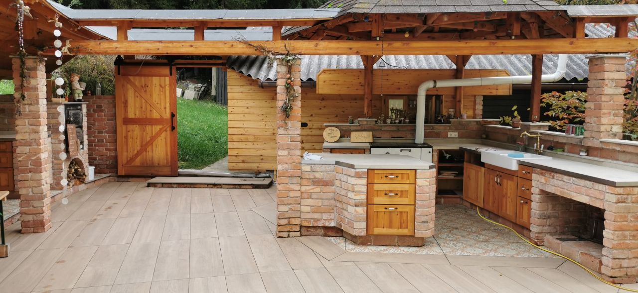 Offene Außenküche aus Ziegelsteinen mit Holzmöbeln und Holzdach und mit Zugang zu einer grünen Wiese.