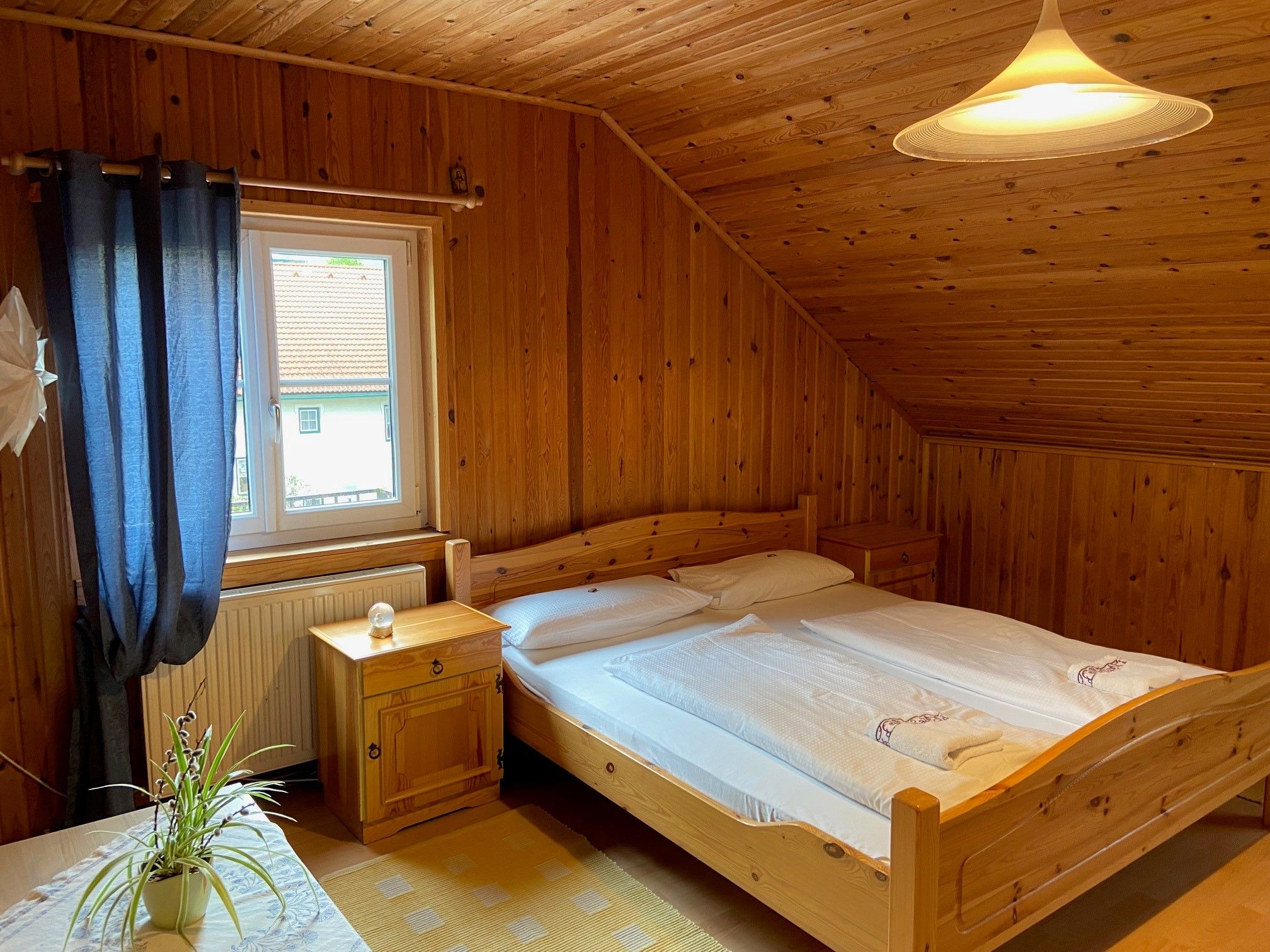 Gemütliches Doppelzimmer mit Holzverkleidung, Doppelbett, Fenster mit blauen Vorhängen und einer Pflanze auf dem Tisch.