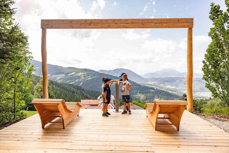 Zwei Kinder stehen auf einer Holzplattform mit Blick auf eine Berglandschaft.