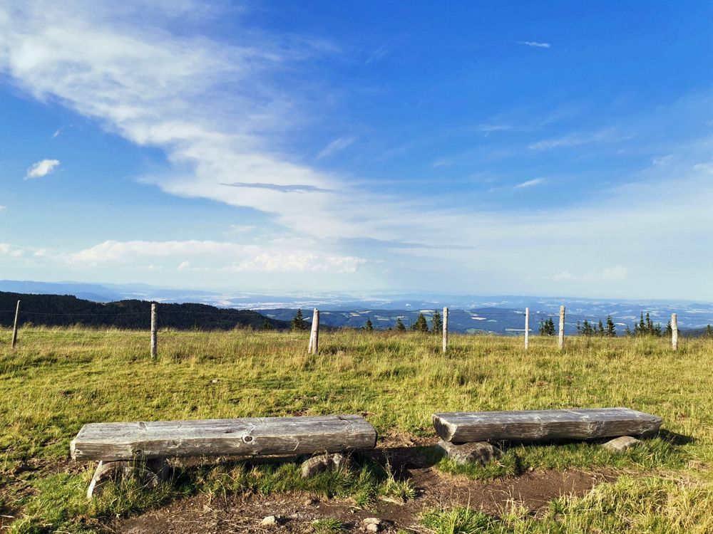 Zwei Holzbänke auf einer Wiese mit weitem Blick über eine Landschaft und blauen Himmel.