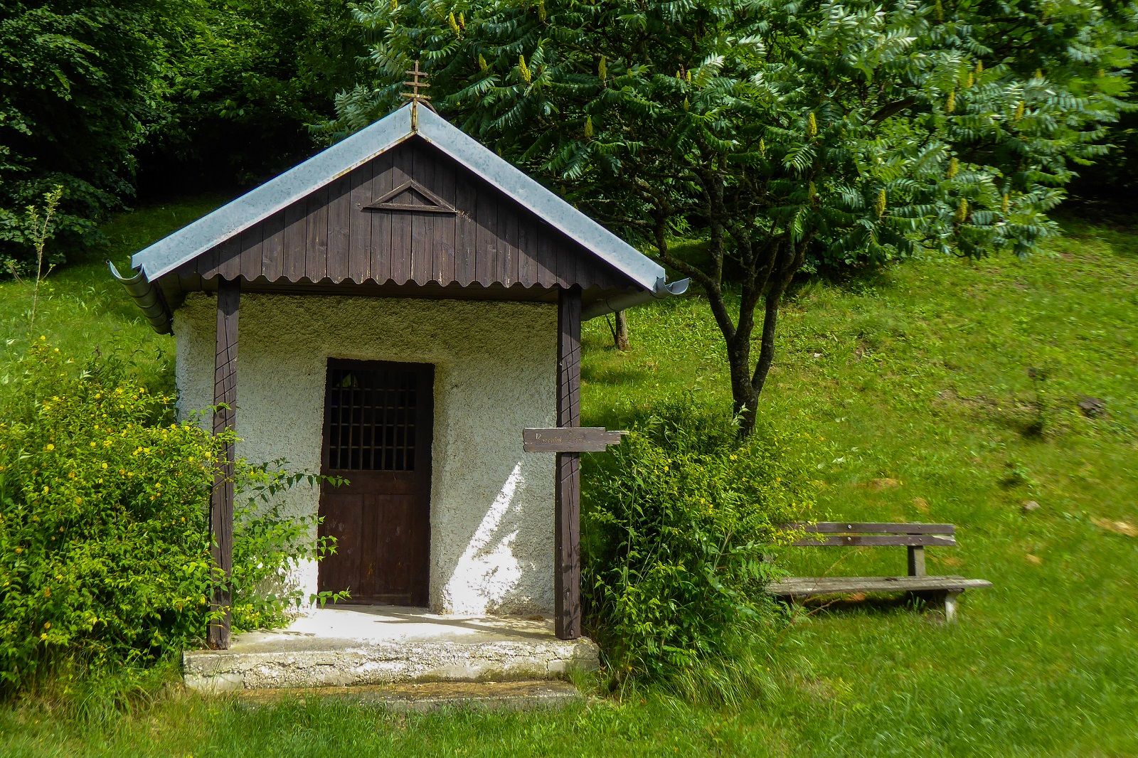 Kleine Kapelle mit Holzdach und Kreuz, umgeben von grüner Vegetation und einer Bank.