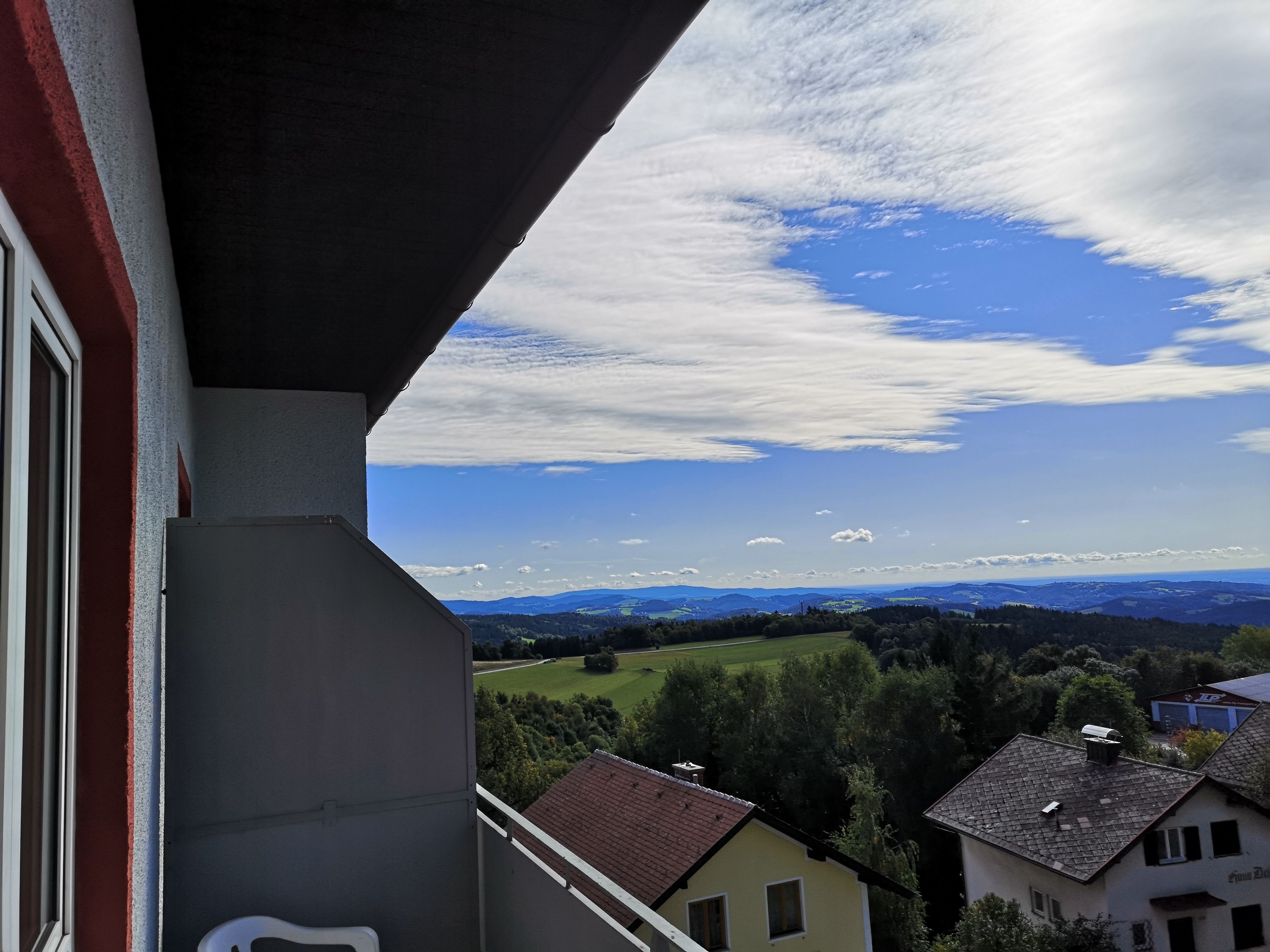 Blick von einem Balkon auf eine Landschaft mit Häusern, Bäumen und Hügeln unter einem blauen Himmel mit Wolken.
