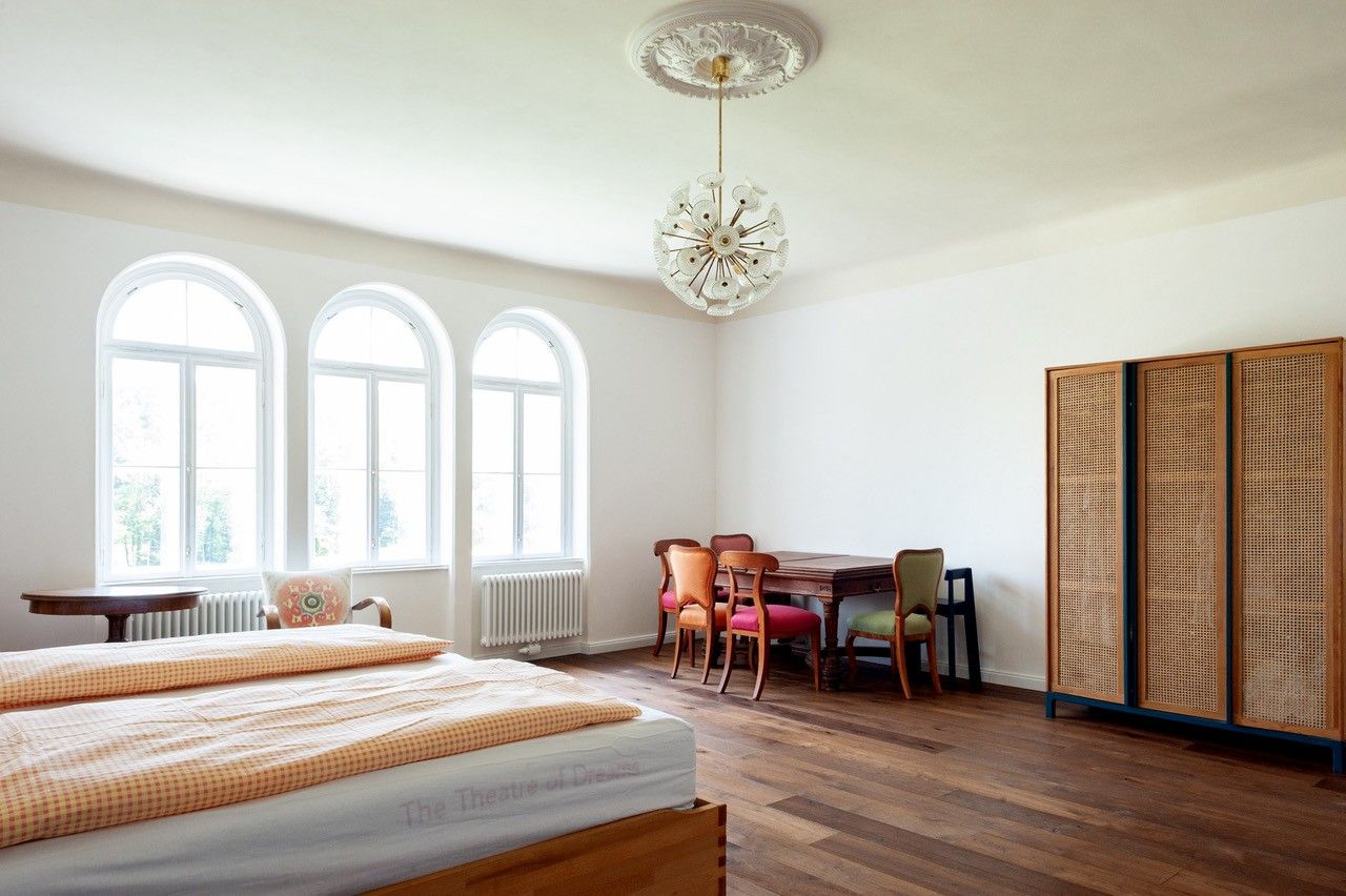 Ein geräumiges Schlafzimmer mit einem großen Bett, einem Tisch mit Stühlen und einem Schrank. Drei große Fenster lassen viel Licht herein.
