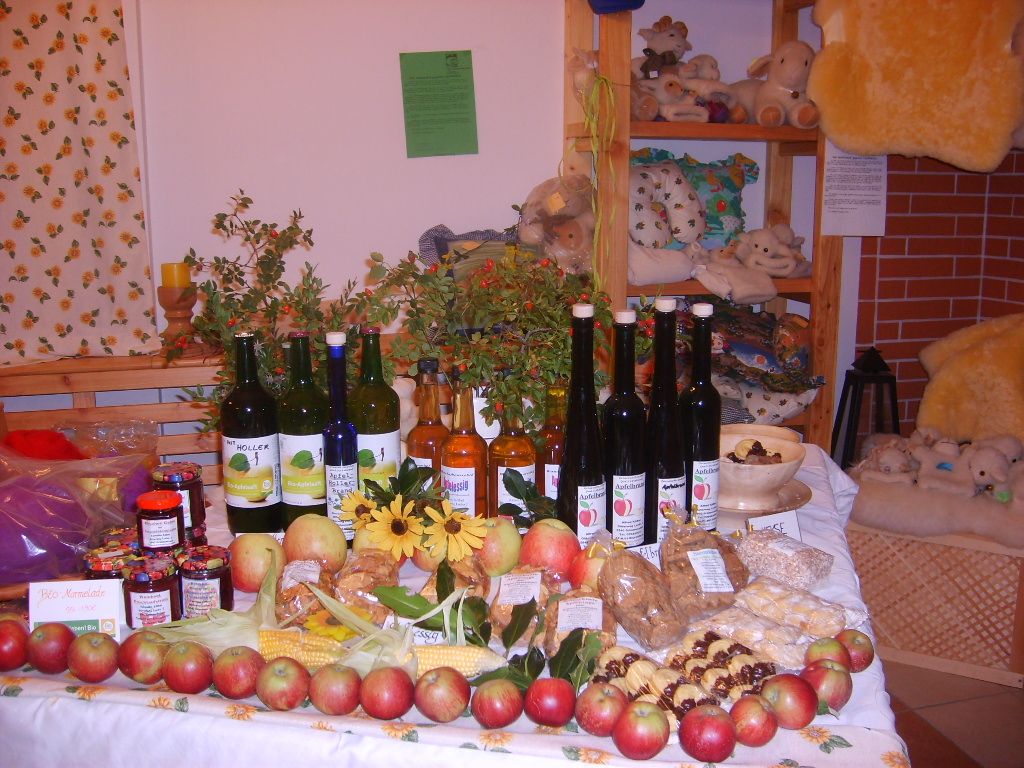 Ein Tisch in einem Hofladen mit verschiedenen Bio-Produkten wie Äpfeln, Marmeladen, Säften und Likören, dekoriert mit Blumen und Pflanzen.