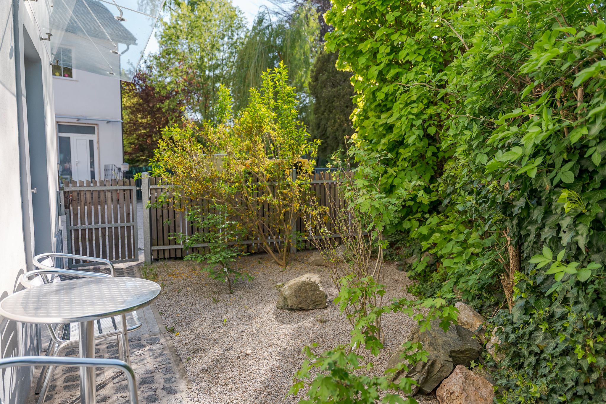 Ein kleiner, gepflegter Garten mit Kiesboden, umgeben von grünen Büschen und Bäumen. Ein runder Metalltisch mit Stühlen steht links im Bild.