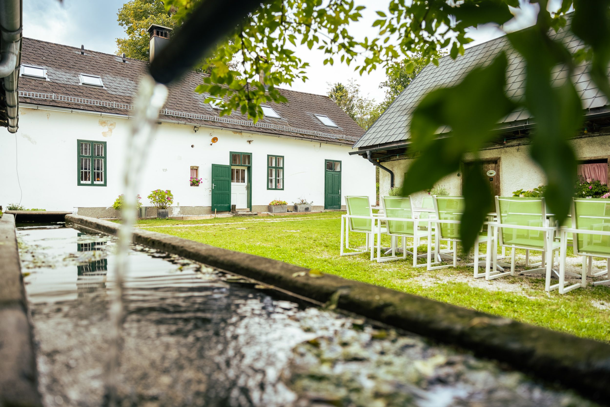 Ein Hof mit weißem Gebäude, grünem Rasen und Gartenmöbeln. Im Vordergrund fließt Wasser in einen Trog.