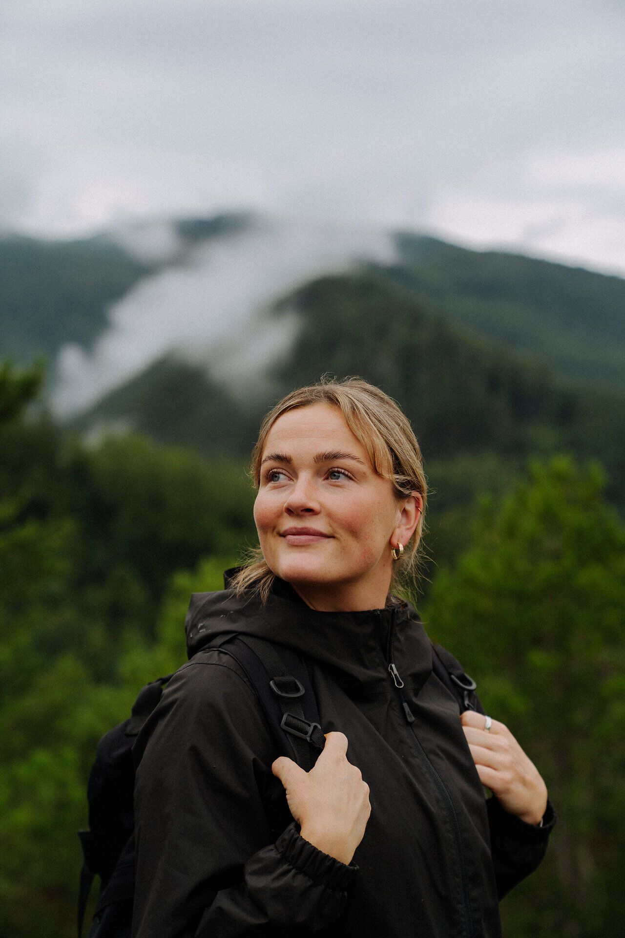 Eine lächelnde Frau in Outdoor-Kleidung mit einem Rucksack blickt zuversichtlich in die Ferne, vor dem Hintergrund einer bewaldeten Berglandschaft mit Nebelschwaden, die den Gipfel umhüllen und eine Stimmung von Abenteuer und Gelassenheit vermitteln.