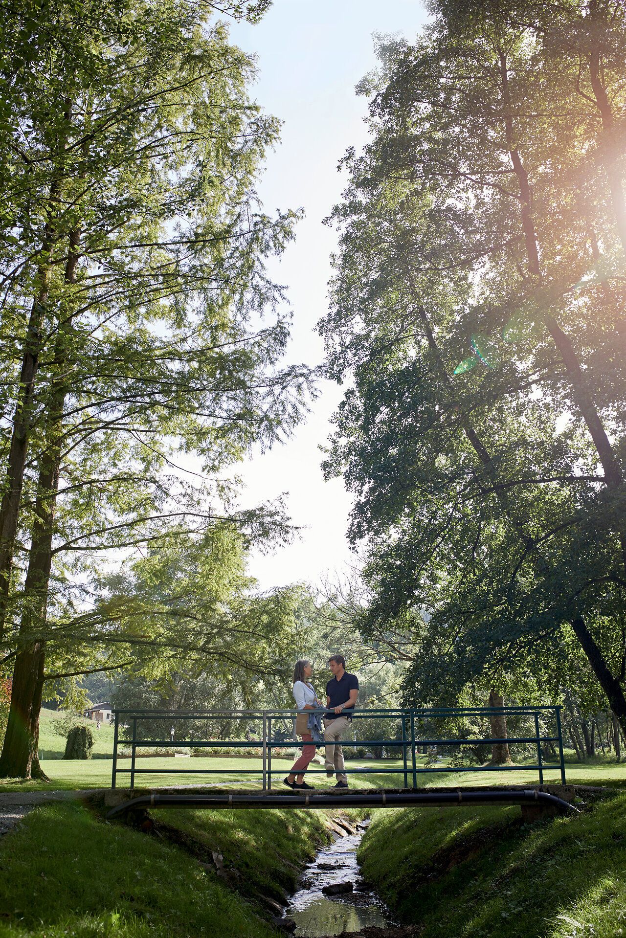 Eine Frau und ein Mann stehen in einem Park auf einer Brücke, umgeben von hohen Bäumen.