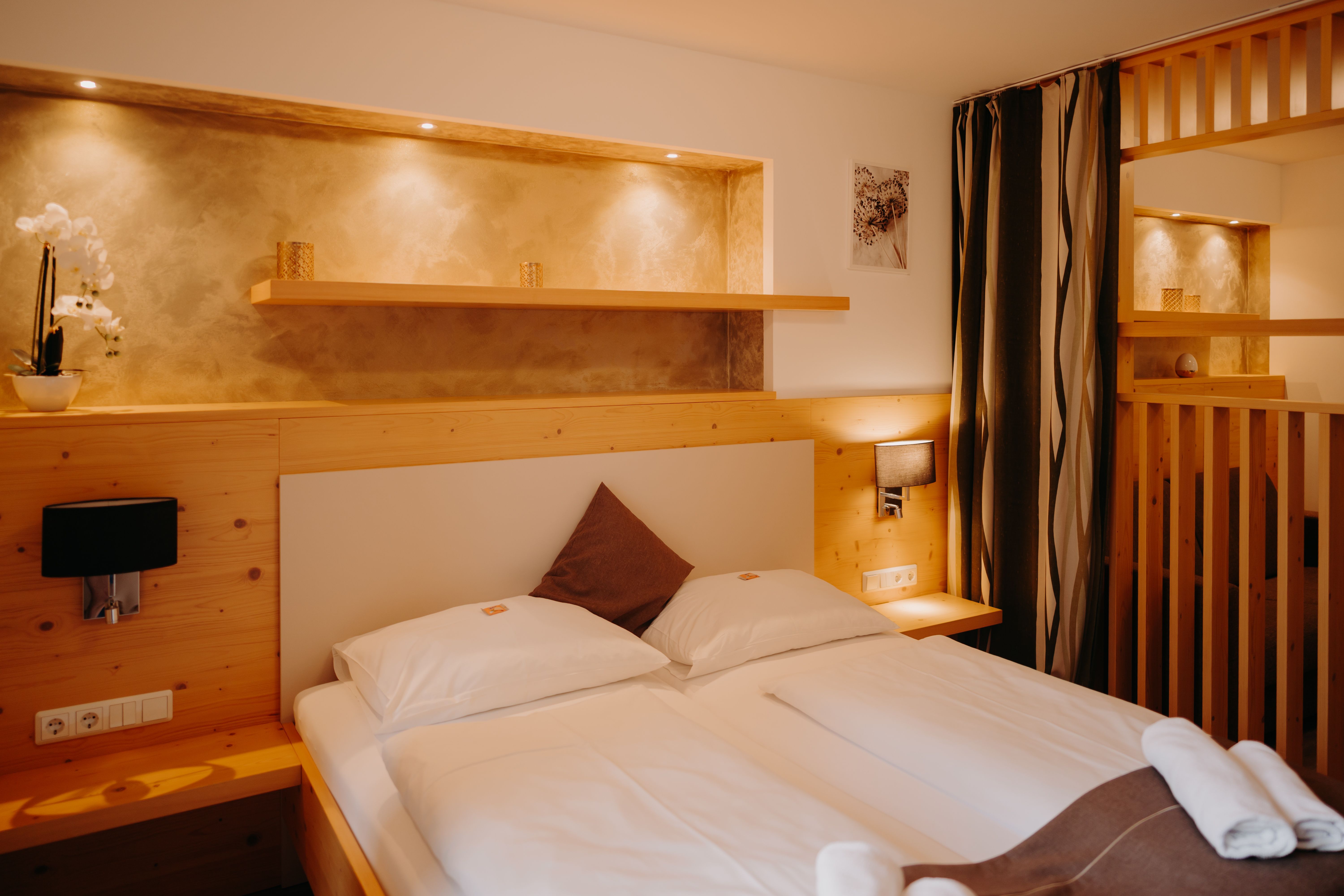 Gemütliches Hotelzimmer mit Doppelbett, heller Holzverkleidung, Raumtrenner aus Holz und mit einem Vorhang. Neben dem Bett sind Steckdosen und Lichtschalter. Über dem Bett eine Nische in der Wand, die beleuchtet und in einem erdigen Goldton gestrichen ist.
