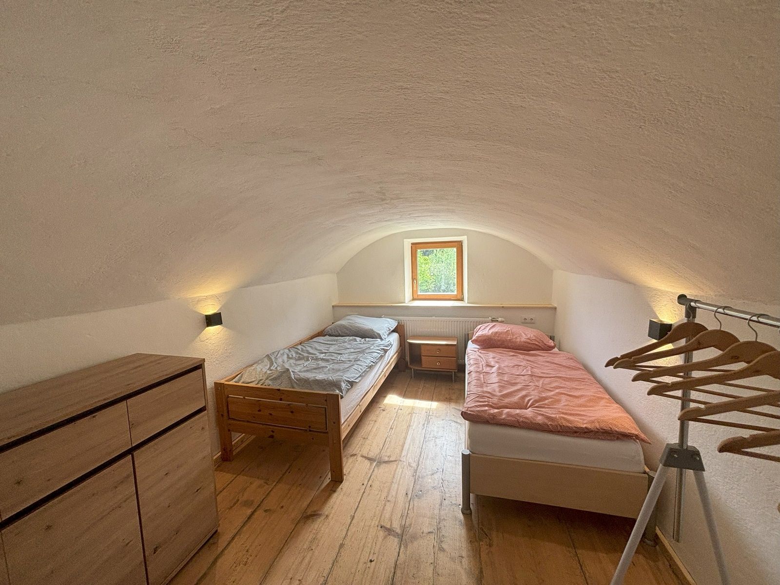 Ein einfaches Doppelzimmer mit zwei Einzelbetten, einer Kommode und einem Kleiderständer. Ein kleines Fenster lässt Tageslicht herein.