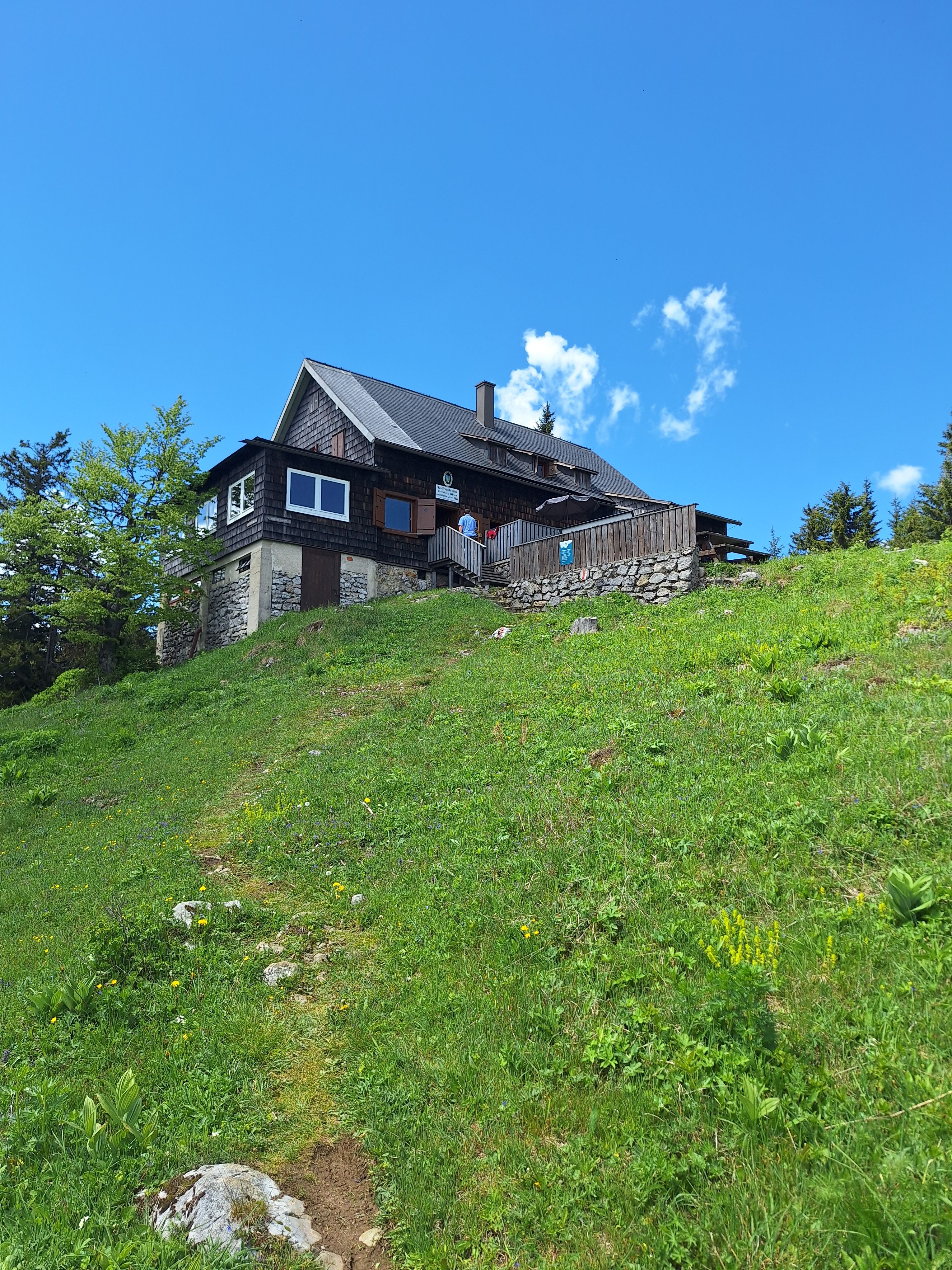 Waldfreundehütte am Obersberg mit Holzverkleidung an einem Hang, davor eine Wiese und blauer Himmel