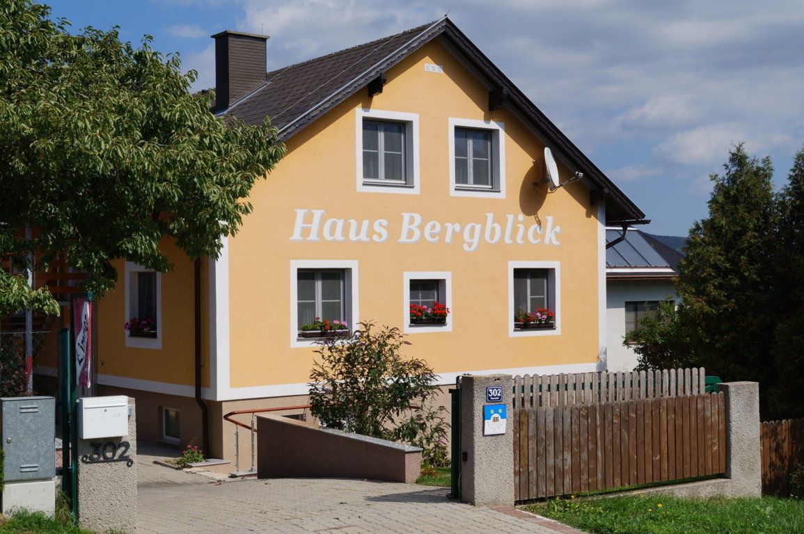Gelbes Haus mit der Aufschrift 'Haus Bergblick', umgeben von Bäumen und einem Holzzaun.